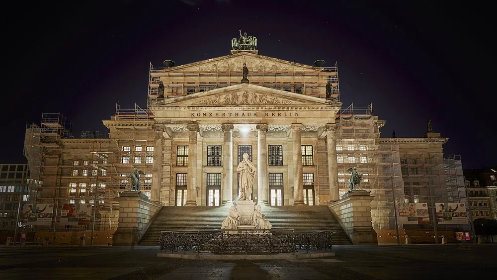 Front view of Konzerthaus Berlin at night, with the Schiller Monument centred before the broad staircase and scaffolding on both sides.