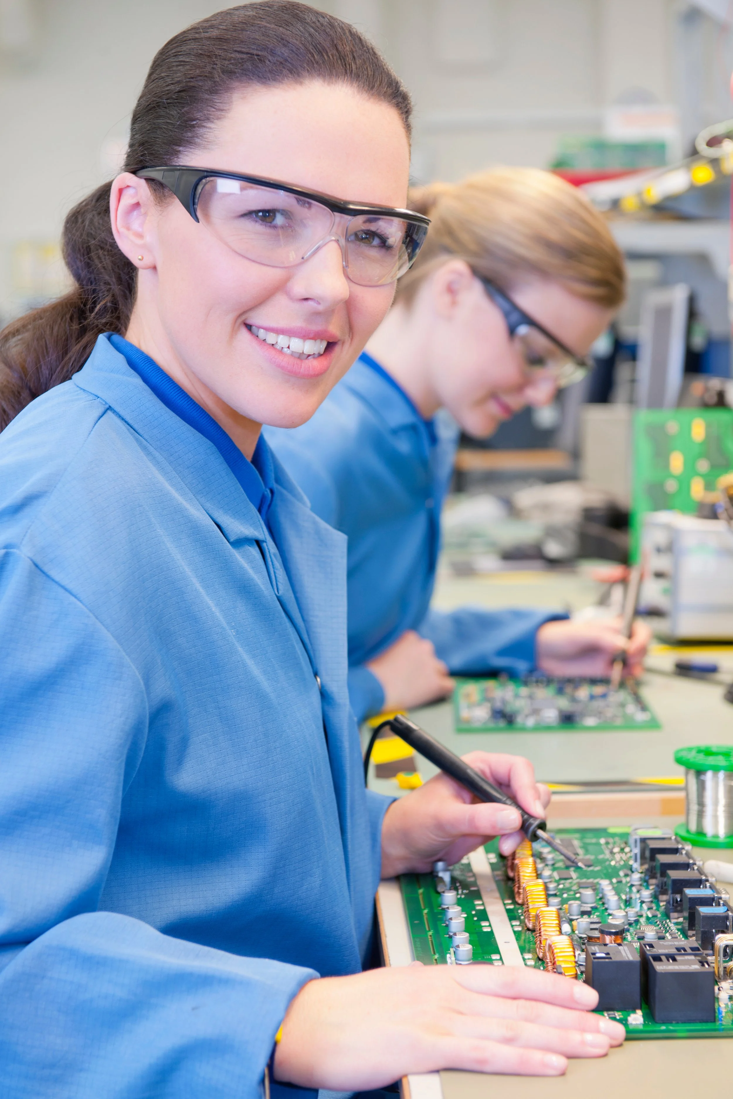 Two women working on electronic circuit boards in a laboratory. The woman in front, smiling and looking at the camera, has dark hair tied back, glasses, and is wearing a blue lab coat. The woman in the background also wears glasses and a blue lab coat, focused on her task.