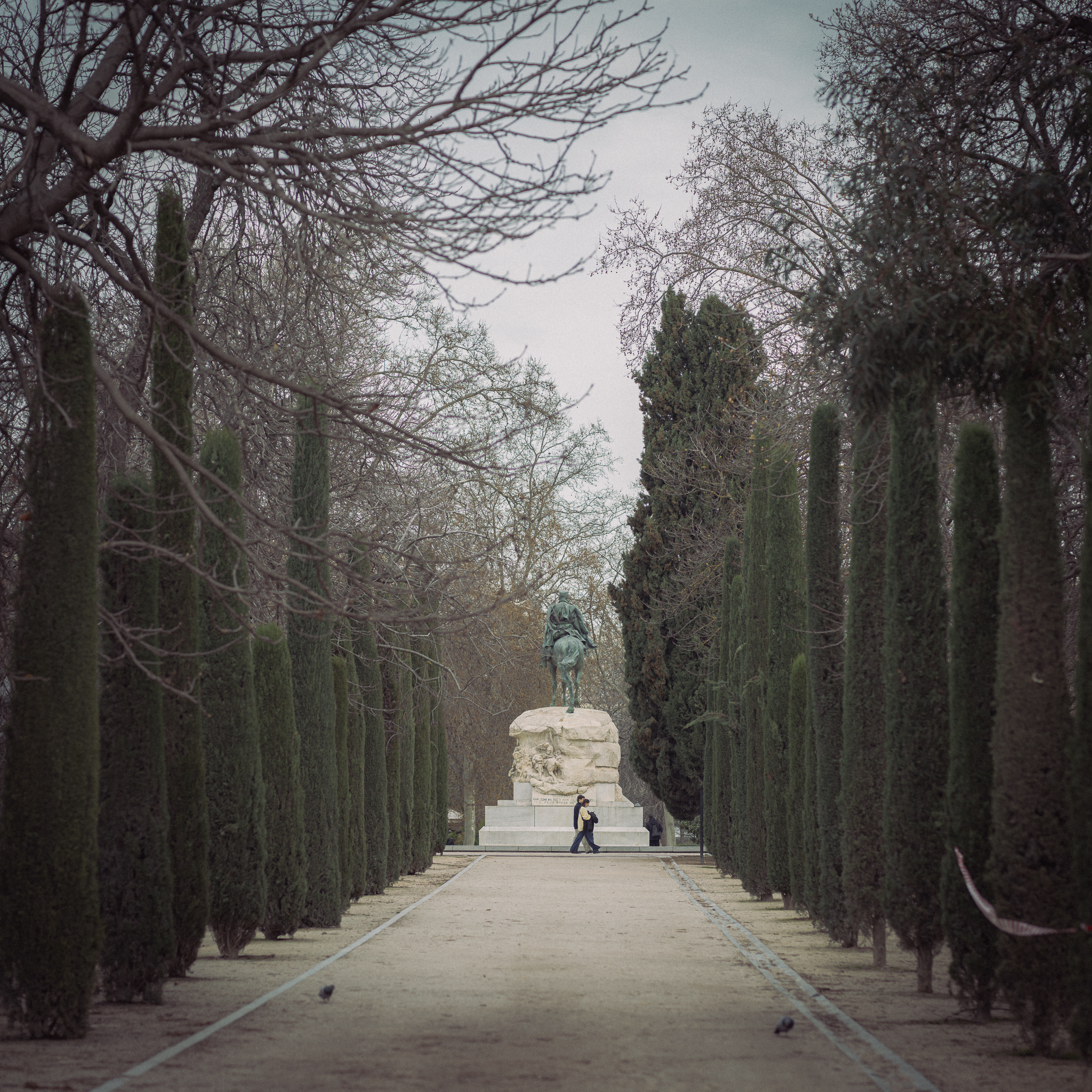 Bronze equestrian monument at the end of a cypress-lined gravel avenue in El Retiro Park, Madrid, on a misty day