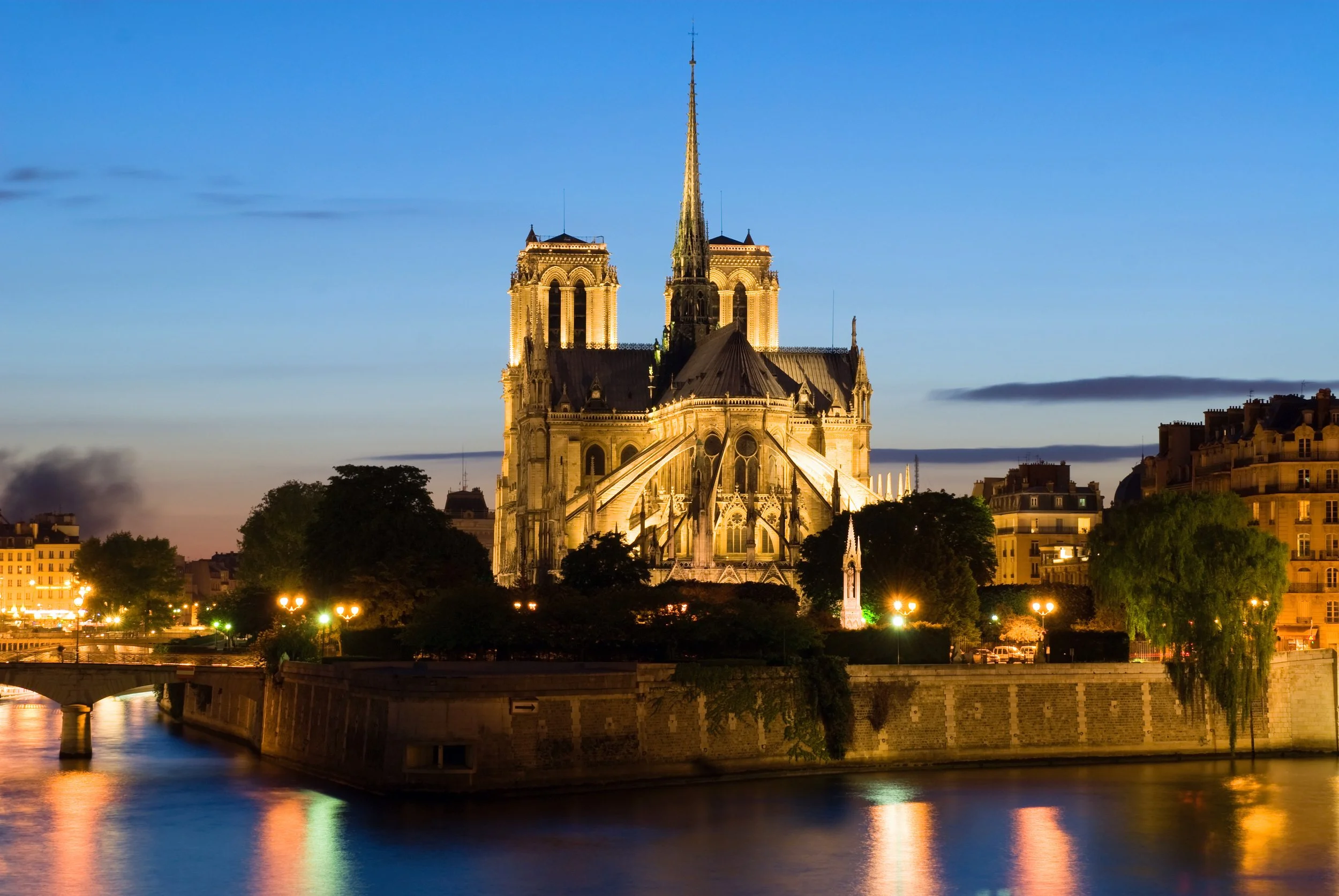 The Notre-Dame Cathedral in Paris at dusk, illuminated with warm lights, with the Seine River and city buildings in the foreground.
