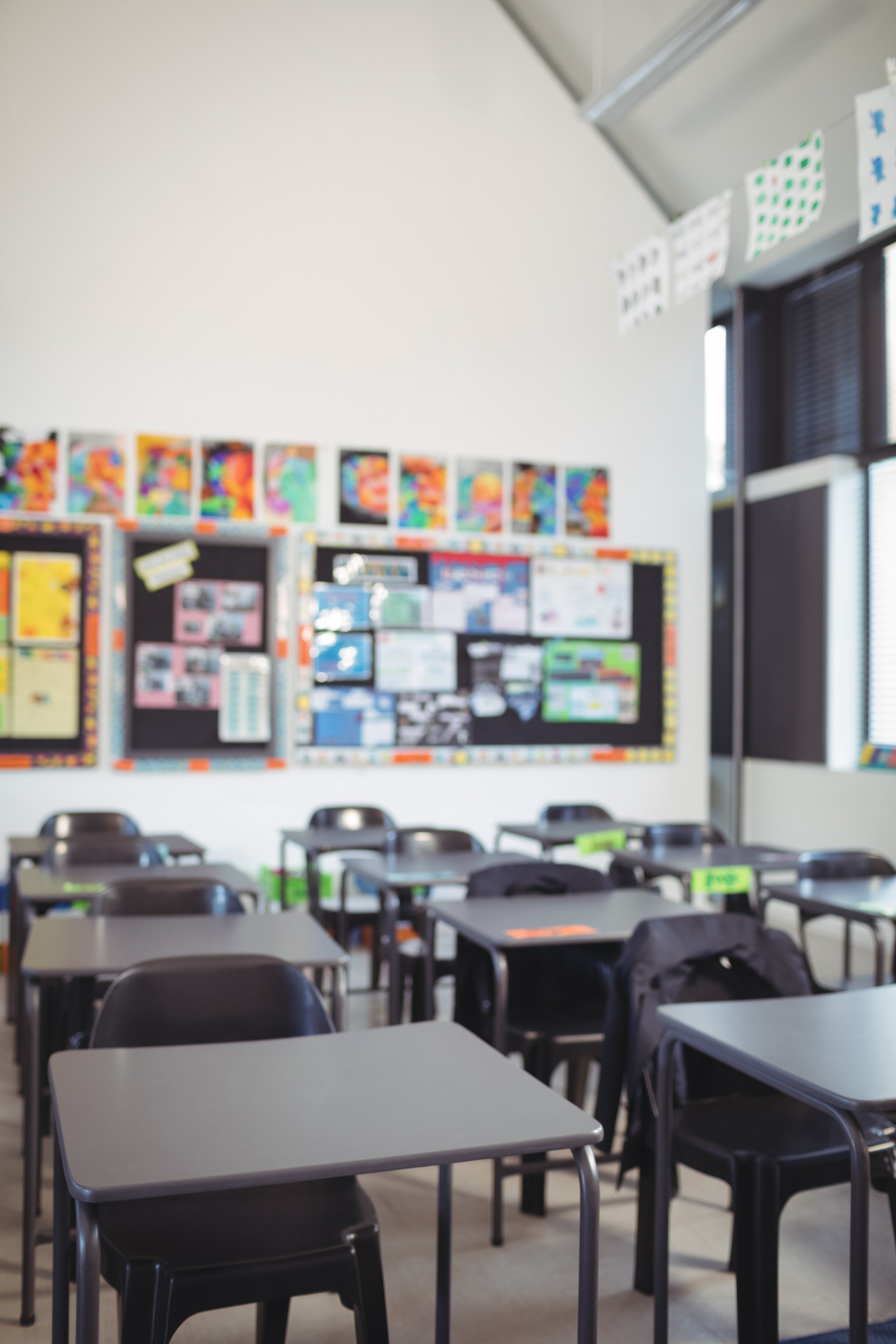 A classroom with empty desks and chairs, a bulletin board with students' artwork and educational posters, and windows with blinds.