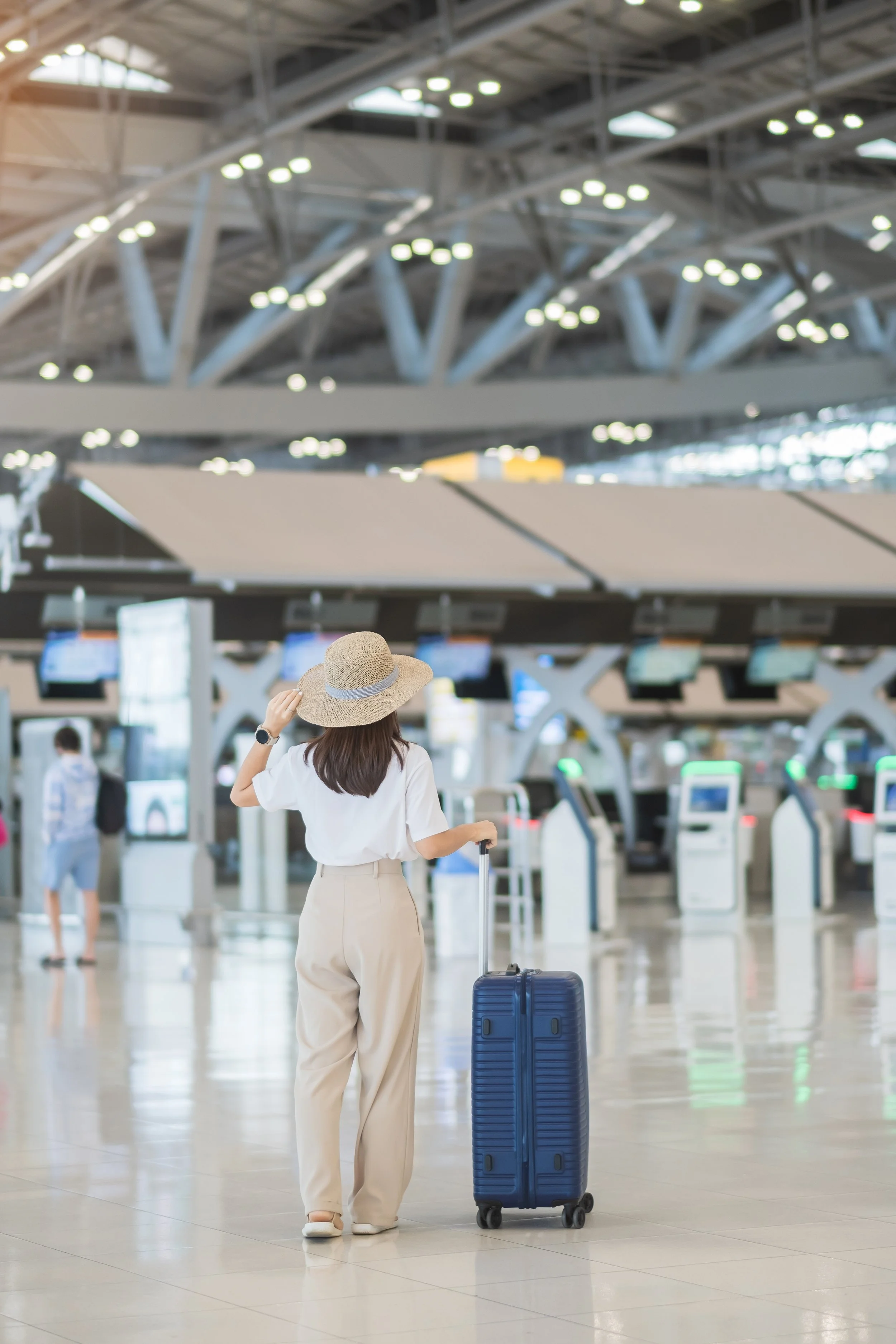 A woman with a suitcase in an airport terminal, facing away, wearing a straw hat, white shirt, and cream-colored pants, with check-in counters and other travelers in the background.