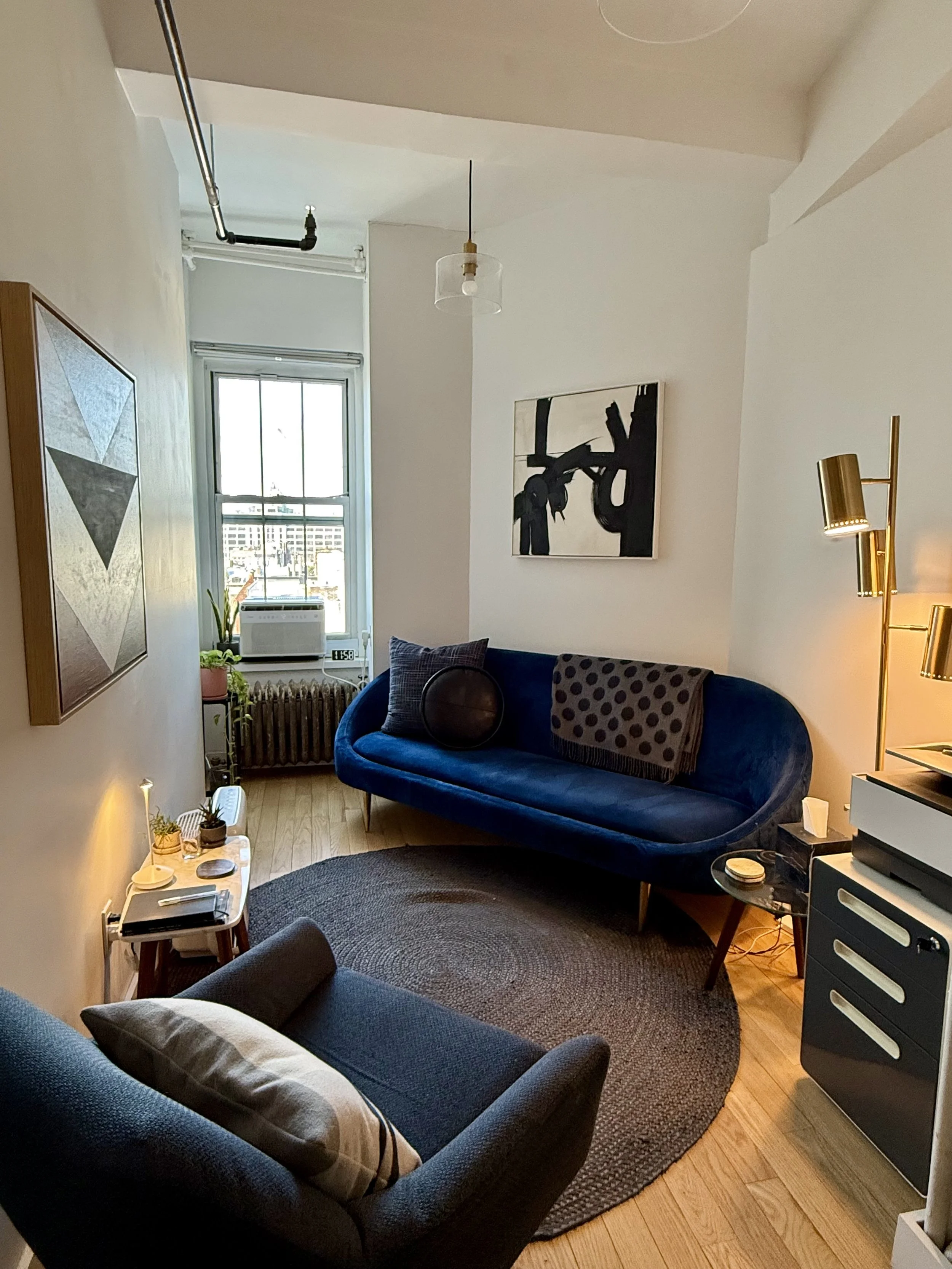 A cozy living room with a navy blue velvet sofa, a gray armchair, a round rug, and wall art, with a large window, potted plants, and warm lighting.
