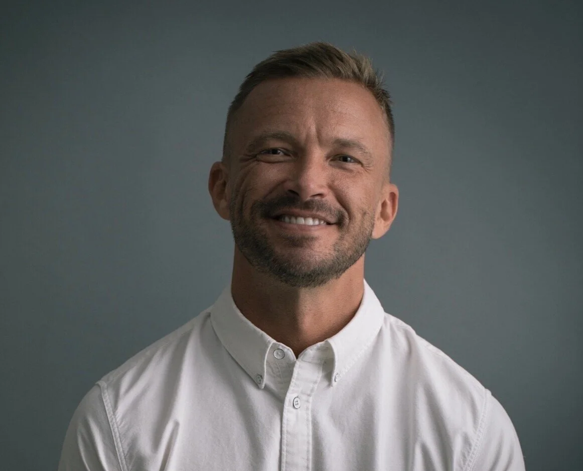 Portrait of a man with short hair, beard, and a white collared shirt, smiling against a gray background.