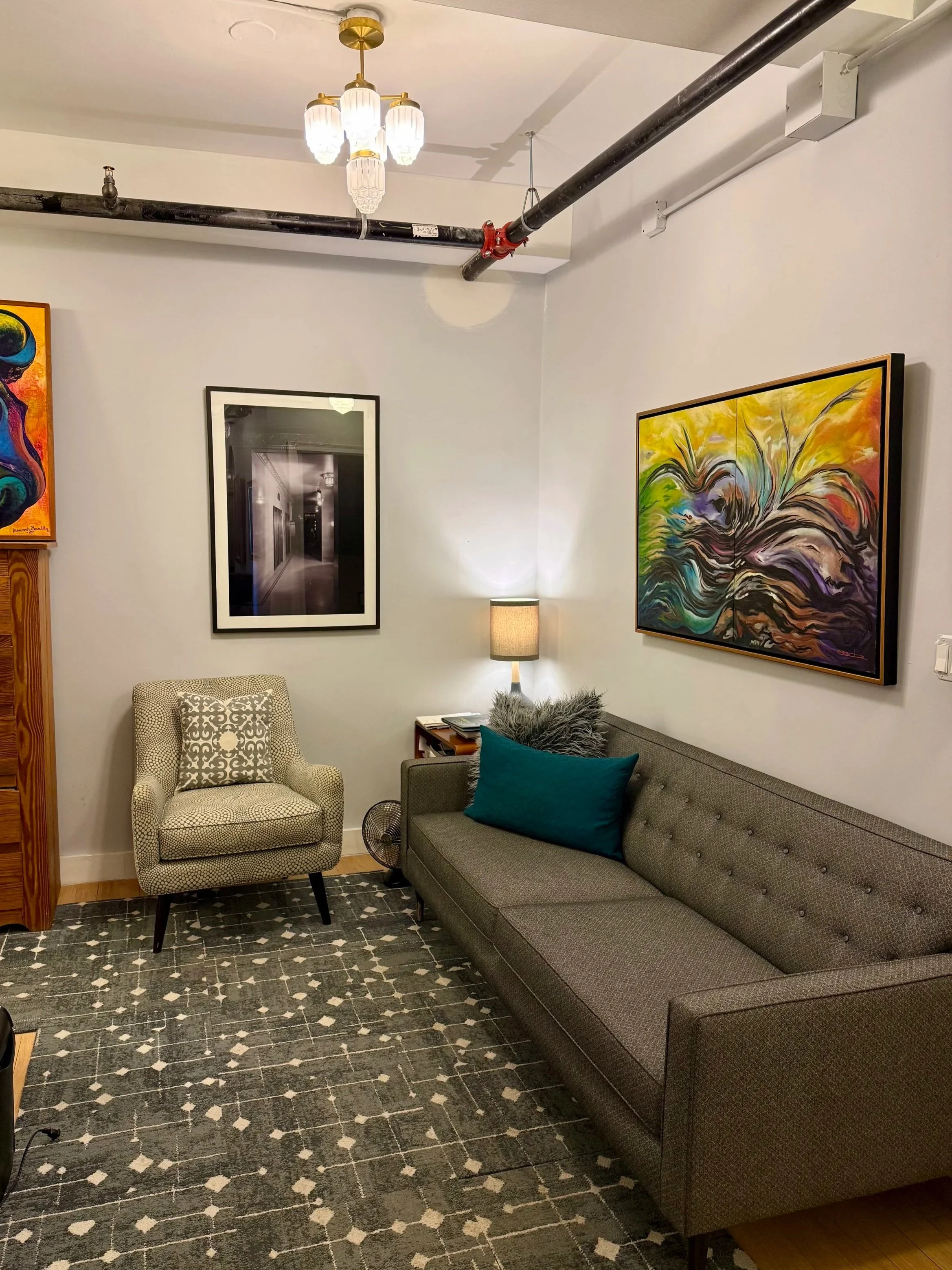 A cozy living room with a gray sofa, patterned armchair, colorful abstract paintings on the wall, a small table with a lamp, a patterned rug, and art decor items.