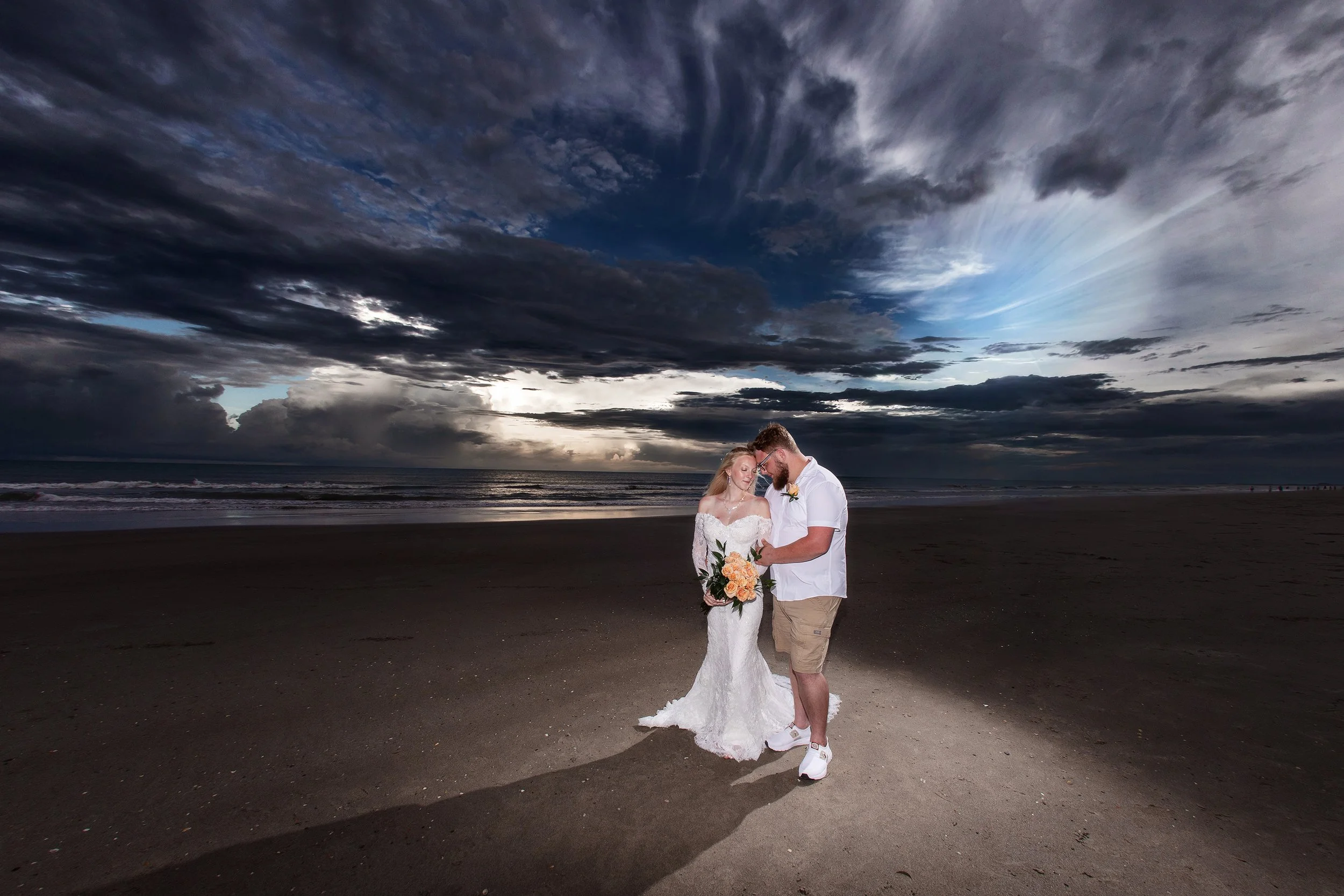 A couple dressed in wedding attire standing on a beach at sunset under a dramatic cloudy sky, with the bride holding a bouquet of flowers.