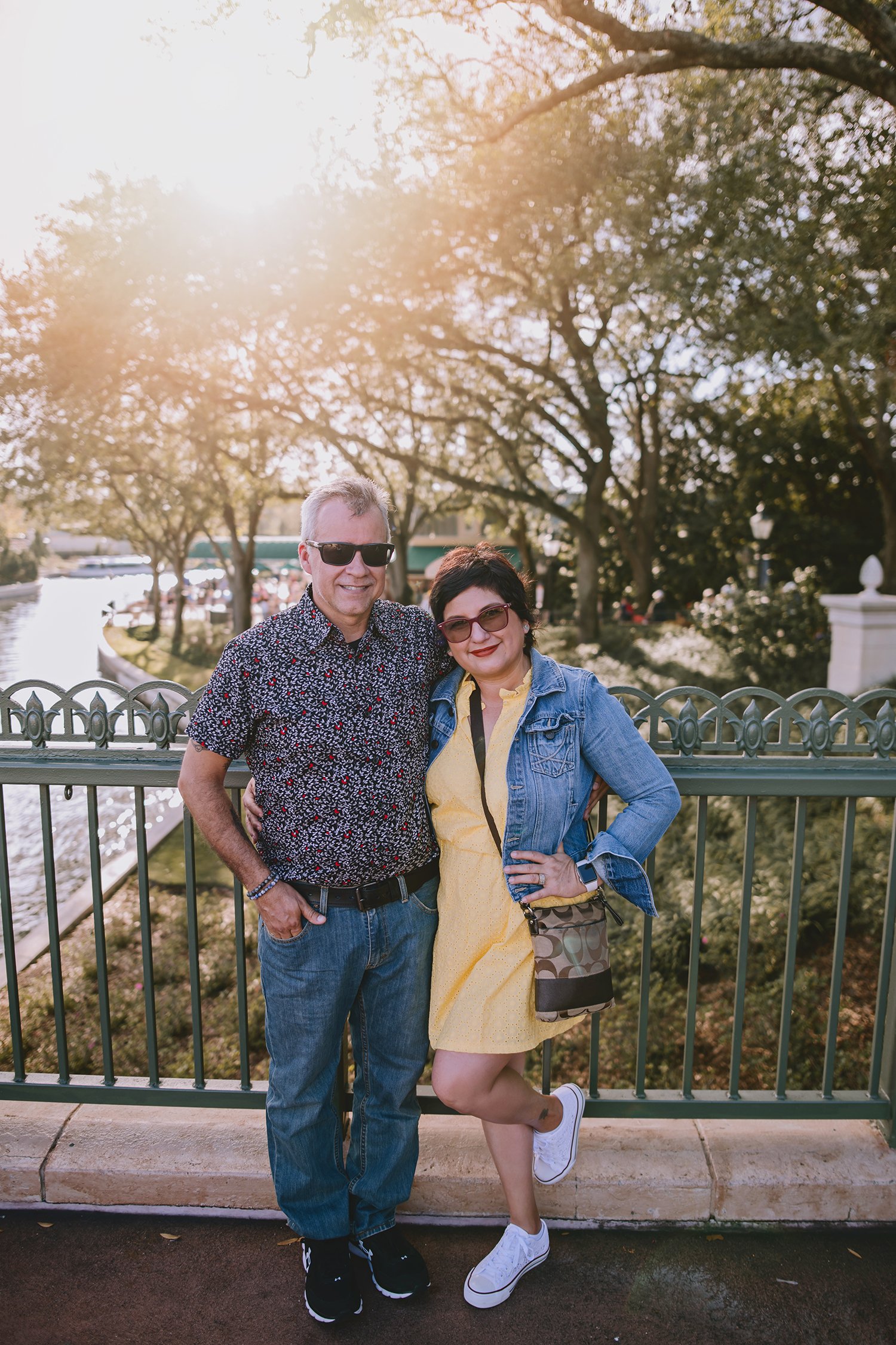 A man and woman standing together outdoors near a river, smiling, with trees and sunlight in the background. The man is wearing sunglasses, a patterned shirt, and jeans, while the woman is wearing sunglasses, a yellow dress, and a denim jacket. They are leaning against a decorative metal fence.