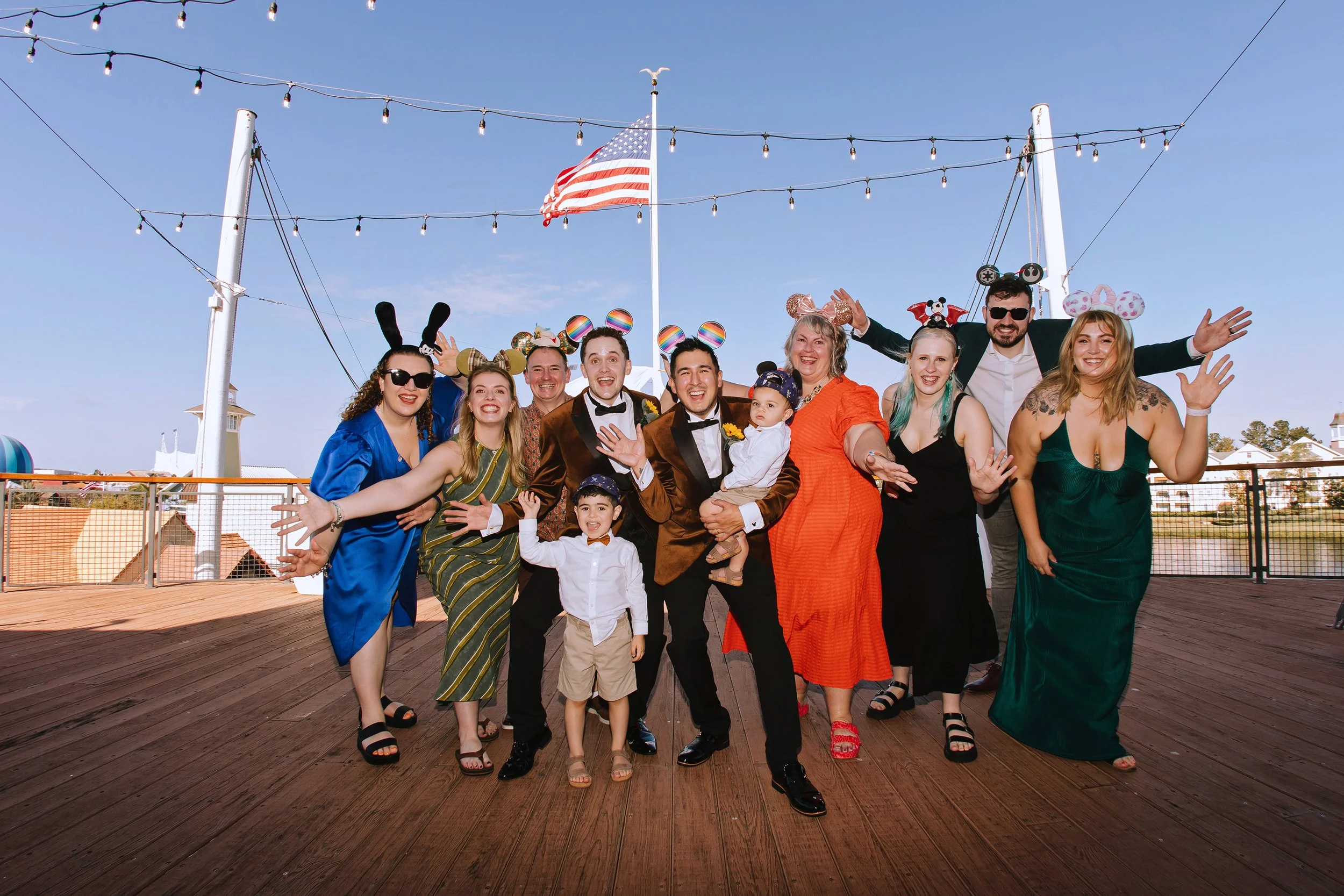 Group of people celebrating on a wooden deck with fairy lights overhead, American flag, and blue sky in the background. They are wearing colorful outfits and fun headbands, smiling and posing for the camera.