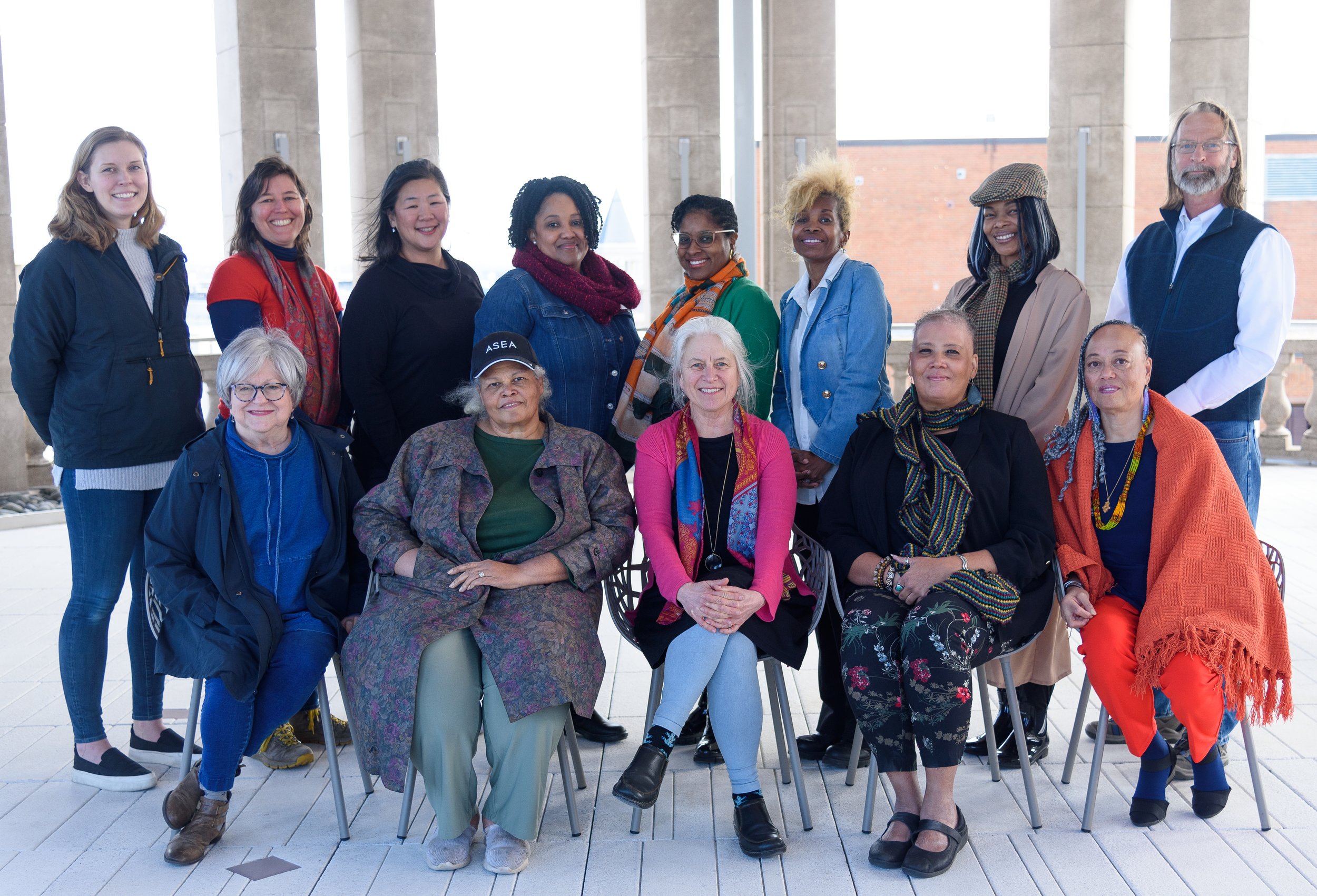 Group of thirteen people, mixed genders, seated and standing, smiling, in casual attire, with a background of pillars and a brick wall.