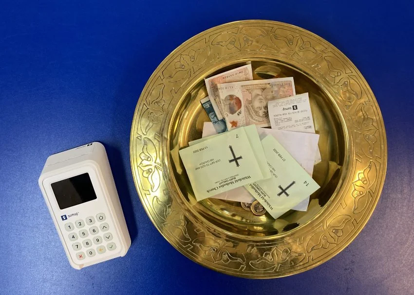 An offering plate with some cash, receipts, and tickets, placed on a blue surface next to a point-of-sale device or card reader.
