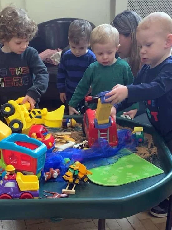 Four children playing with various colorful toy construction vehicles and tools on a large black table, with a woman assisting in the background.