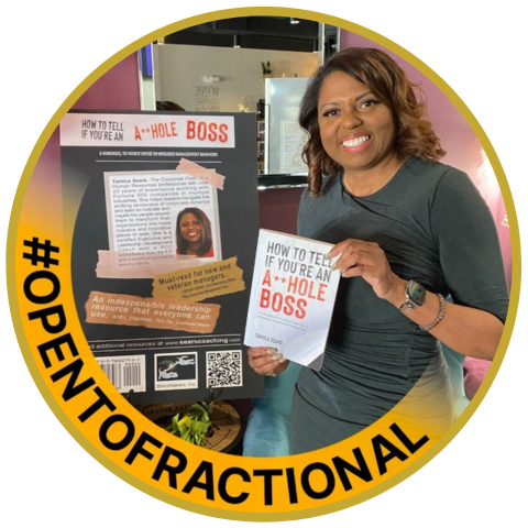 A woman smiling and holding a flyer that reads 'How to Tell if You Are an A-Hole Boss' standing next to a display board with similar text, in an indoor setting.