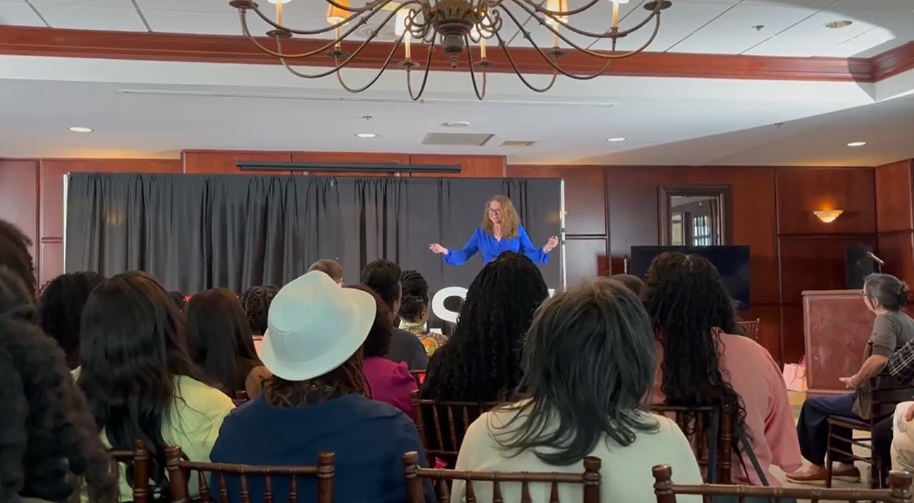 A woman with curly hair in a blue blouse giving a TEDx Talk on a stage in front of an audience in a banquet room.