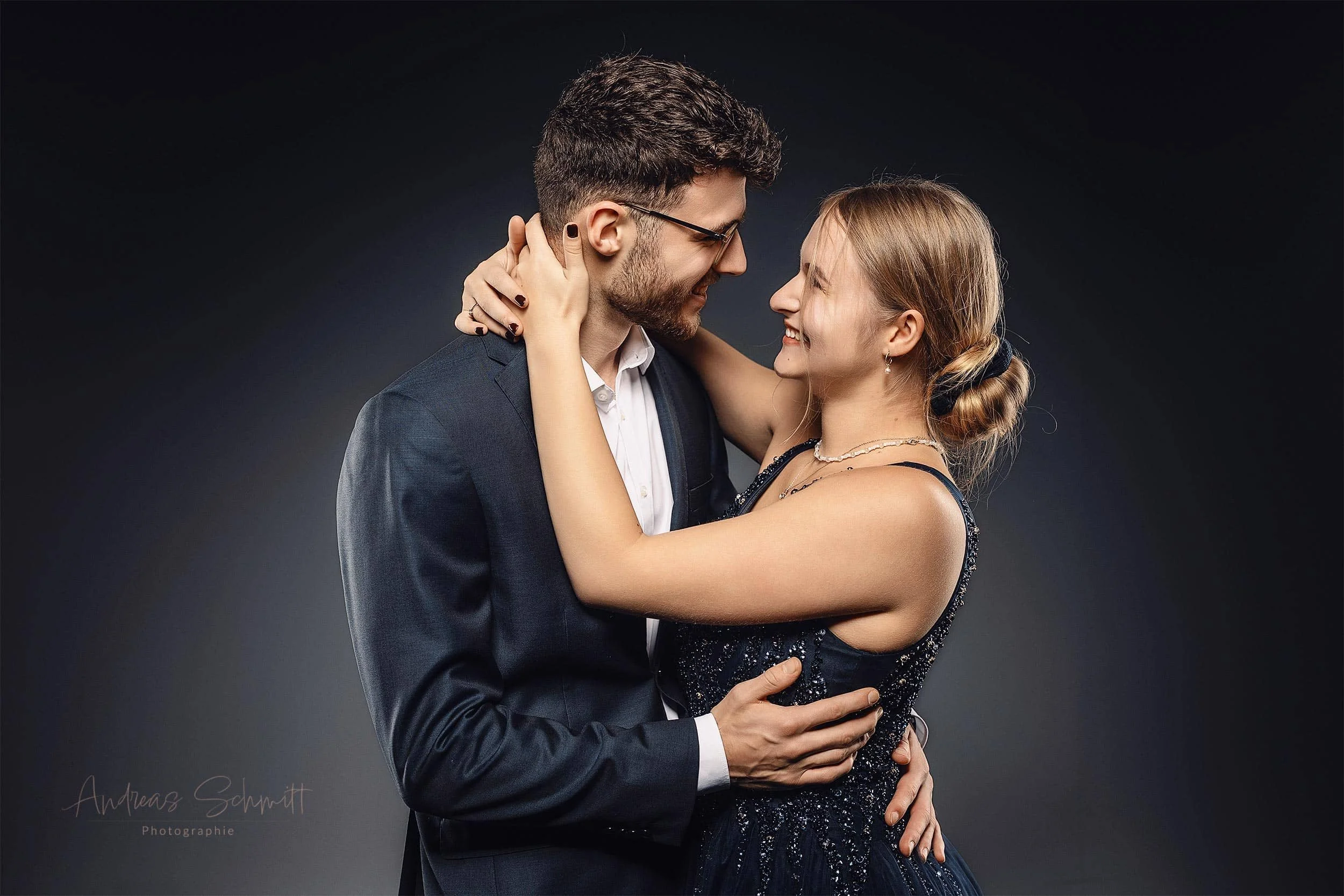 Ein Mann und eine Frau umarmen sich liebevoll, beide lächeln sich an. Der Mann trägt einen dunklen Anzug und die Frau ein schwarzes Abendkleid mit Glitzern.