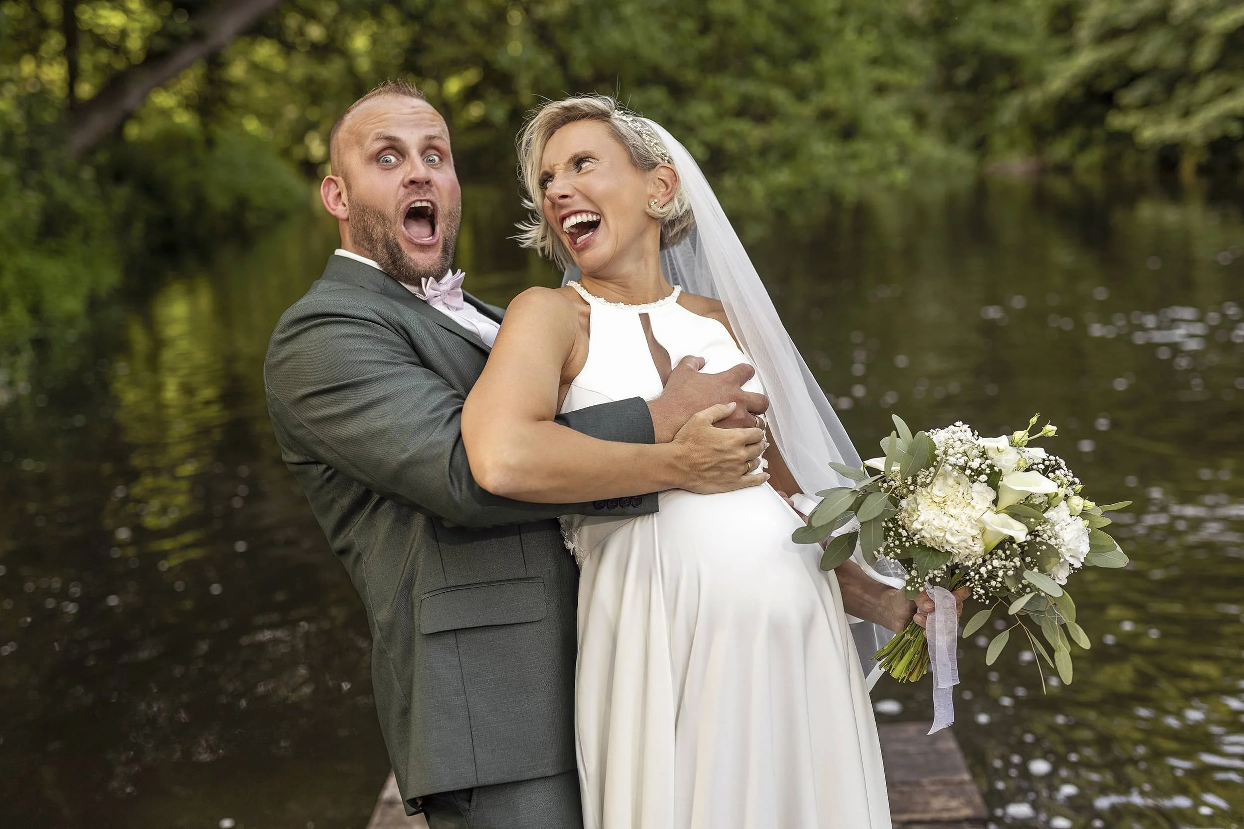 Ein Mann in Anzug und eine Frau in einem weißen Hochzeitskleid lachen und posieren vor einem Wasserhintergrund.