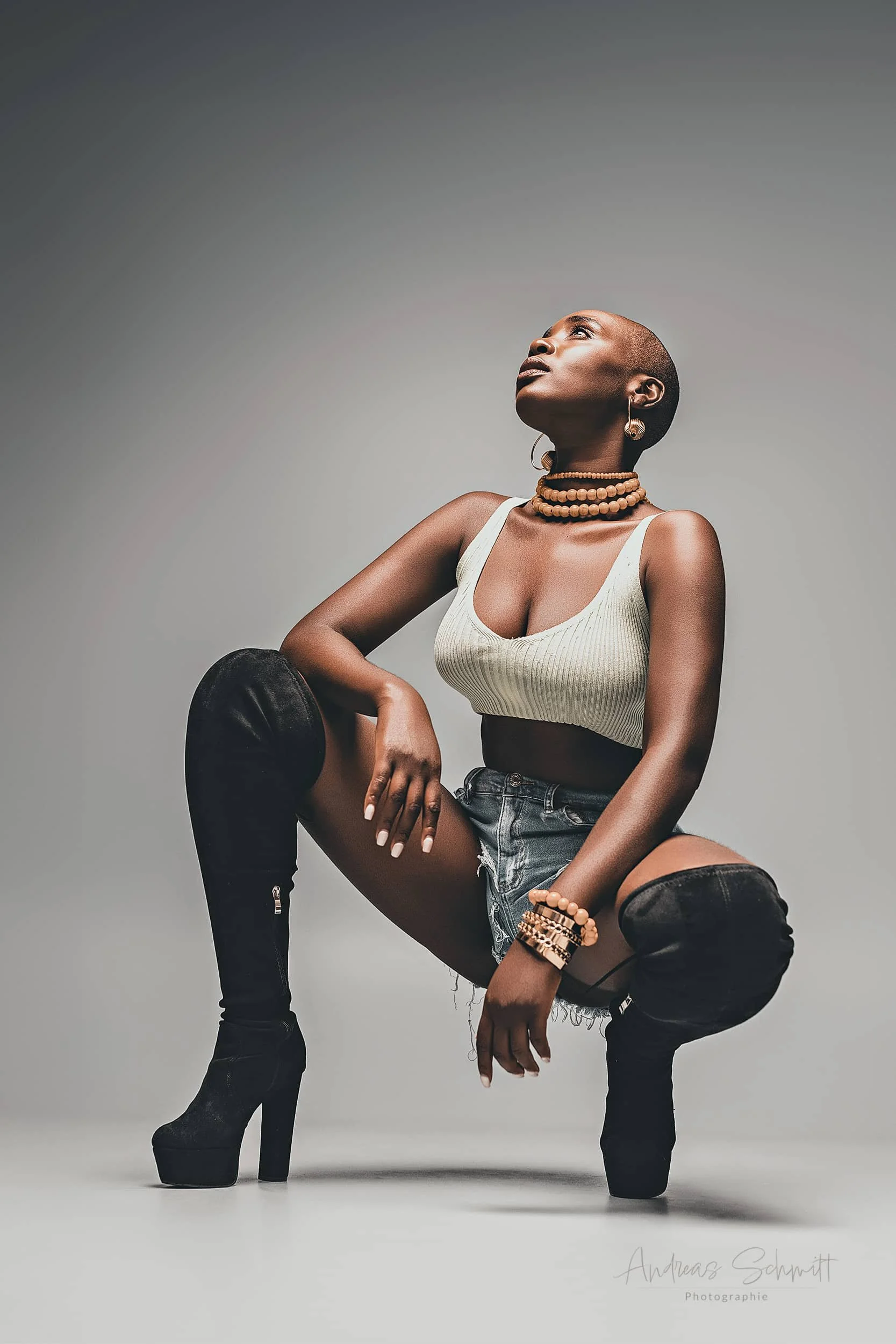 Eine asiatische Frau sitzt auf dem Boden, trägt schwarze Overknee-Stiefel mit hohen Absätzen, eine weiße Crop-Top, kurze Denim-Shorts und Schmuck wie Halsketten, Armbänder und Ohrringe. Sie blickt nach oben gegen einen grauen Hintergrund.