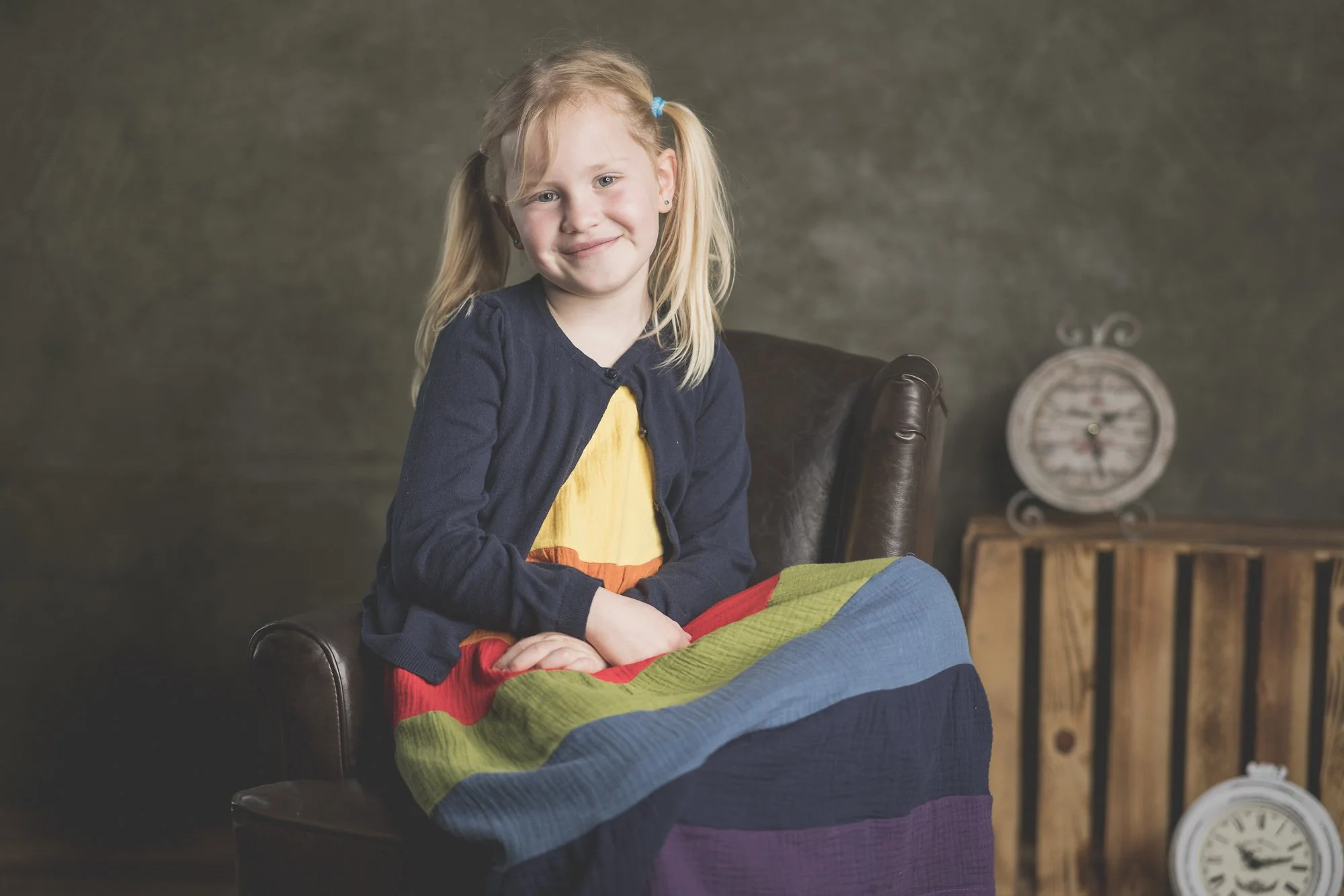 Ein Mädchen mit blonden Haaren sitzt auf einem Ledersessel, trägt einen bunten Rock und einen dunklen Cardigan. Im Hintergrund steht eine Holzbox mit einer dekorativen Uhr darauf.