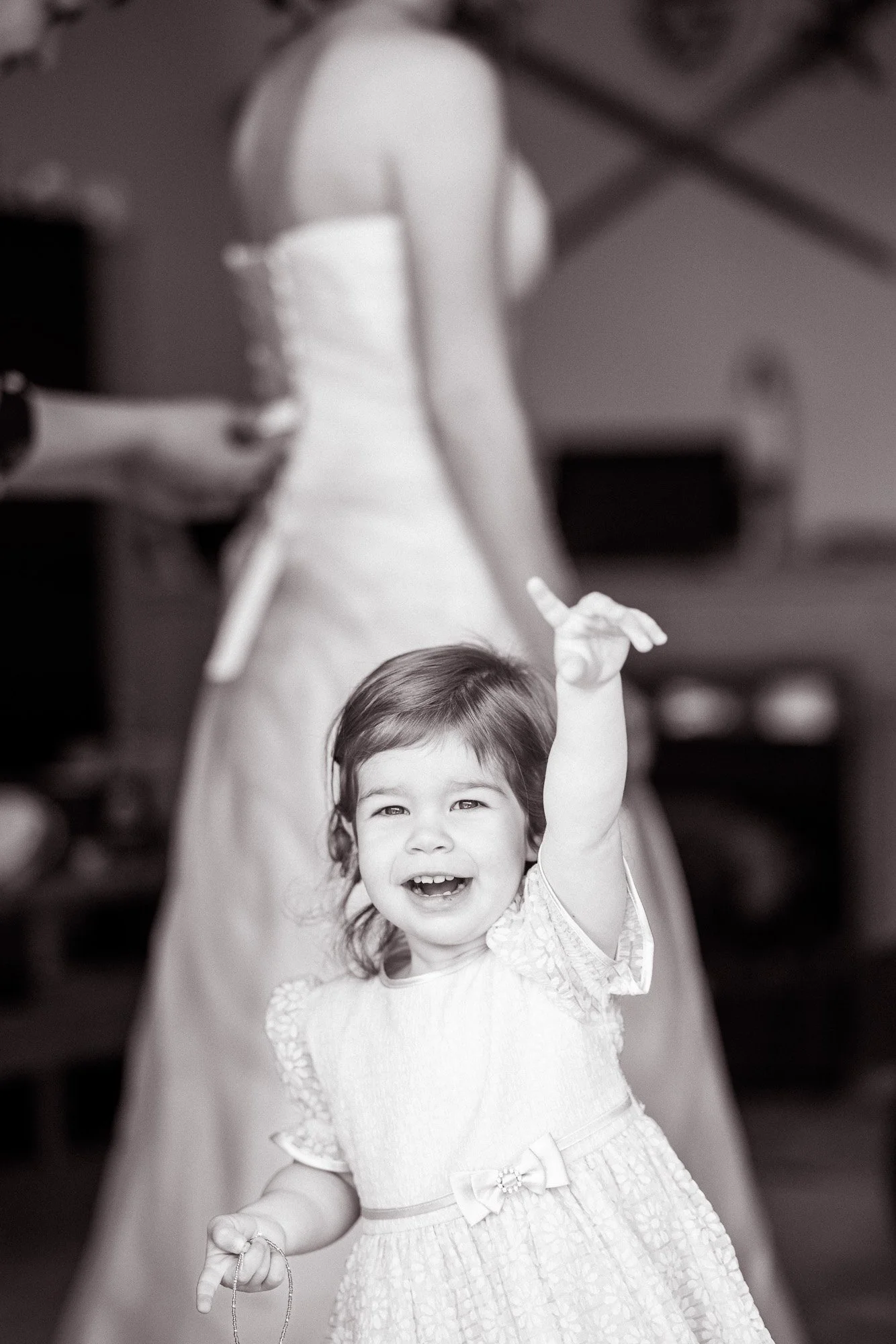 Ein lachendes kleines Mädchen mit braunen Haaren in einem Kleid, das in einem Innenraum steht, während eine ältere Person im Hintergrund steht.