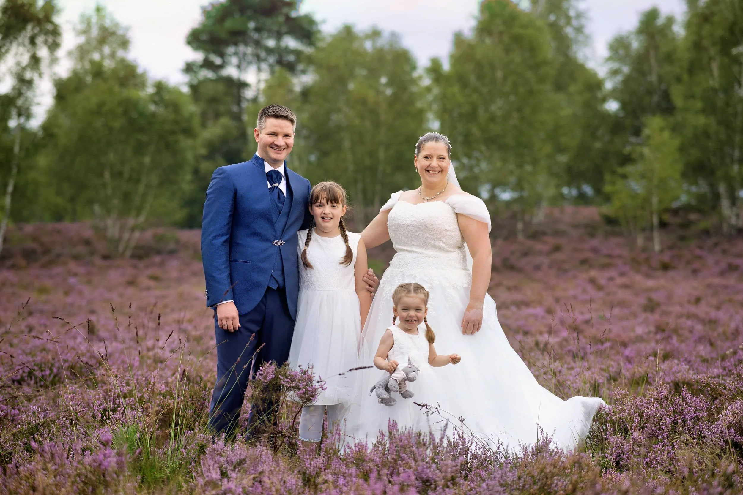 Hochzeitspaar mit zwei jungen Mädchen in einem blühenden Heidefeld, im Hintergrund Bäume und Himmel.