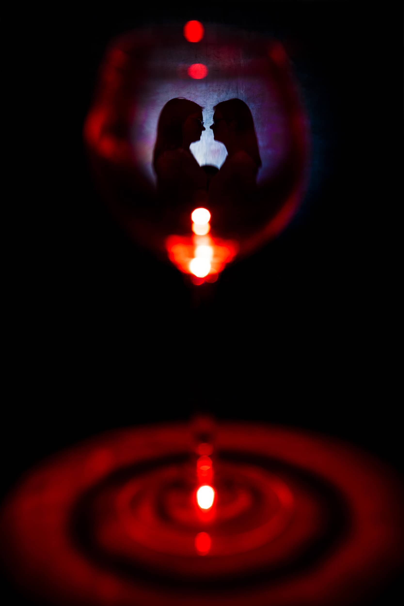 Silhouetten zweier Frauen, die sich gegenüberstehen, sichtbar durch eine rote Linse vor einer dunklen Hintergrund, mit Reflexion im Wasser.