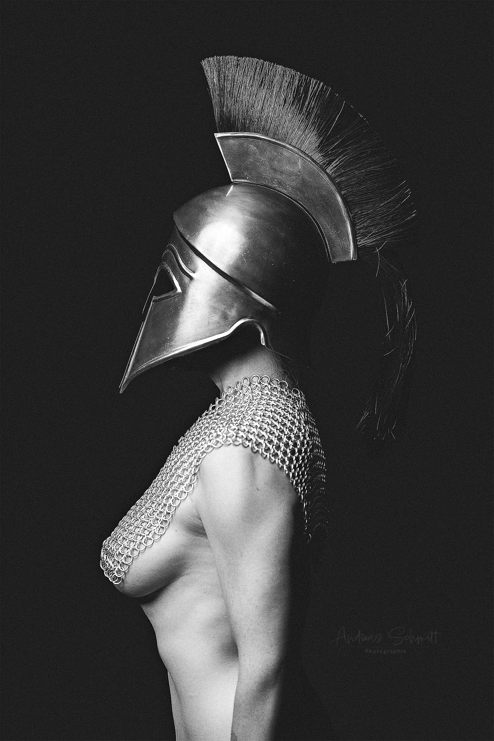 Schwarz-Weiß-Fotografie einer Person mit einer antiken römischen Legionärsmaske, einem Metal-Kettenpanzer und nacktem Oberkörper, gegen einen dunklen Hintergrund.