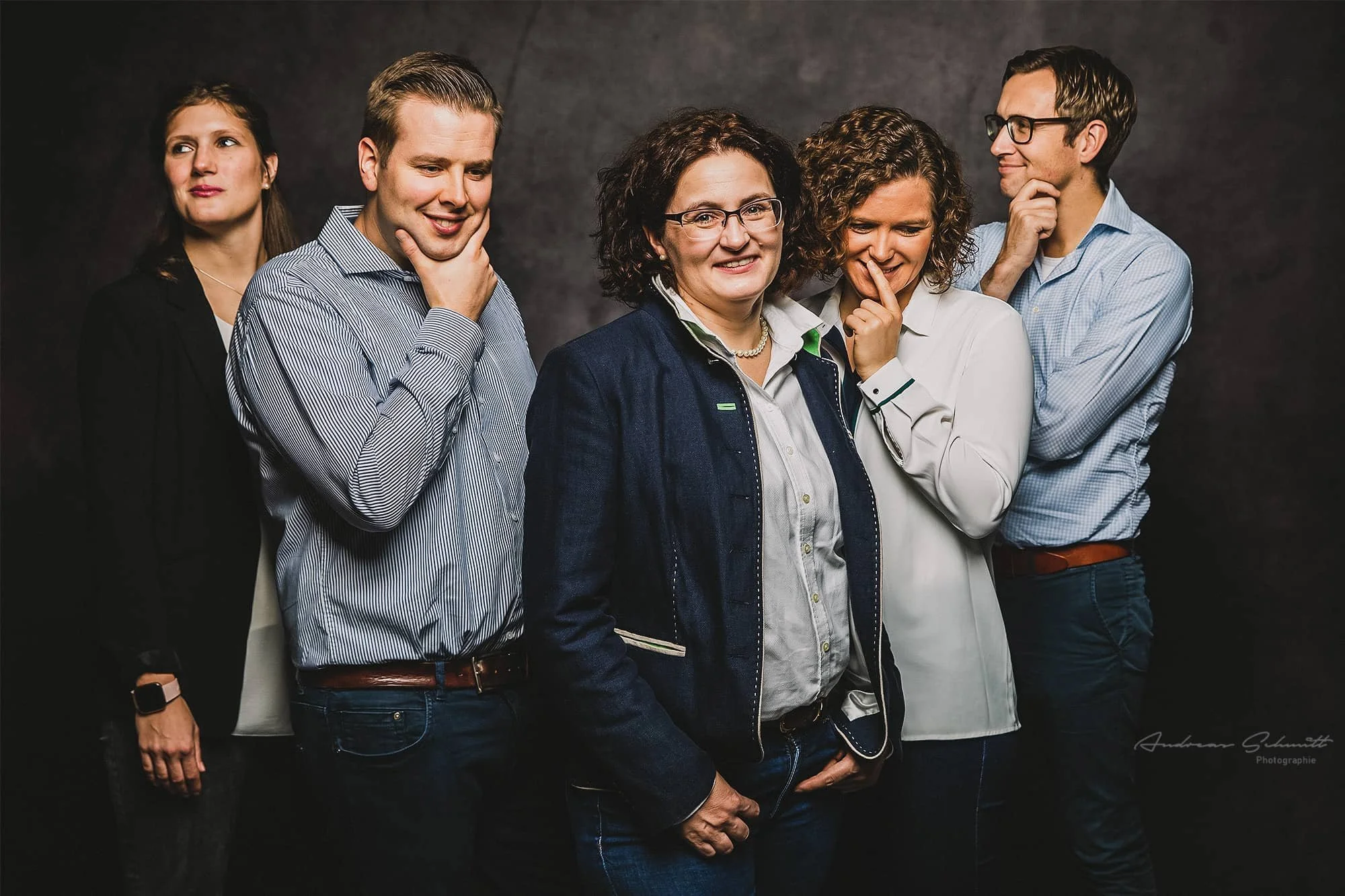 Fünf Menschen posieren vor einem dunklen Hintergrund, lachend und nachdenklich, in Business-Kleidung.