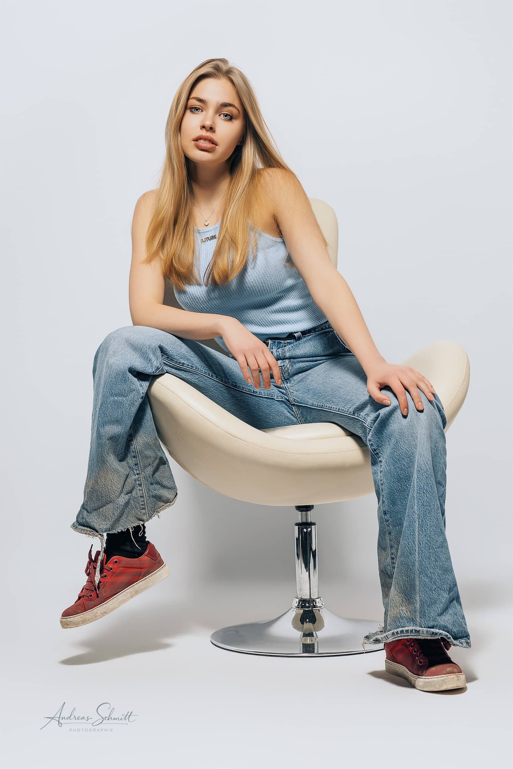 Eine junge Frau mit langen blonden Haaren sitzt auf einem weißen, modernen Stuhl, trägt eine blaue ärmellose Oberteil und lose Jeans, und schaut in die Kamera