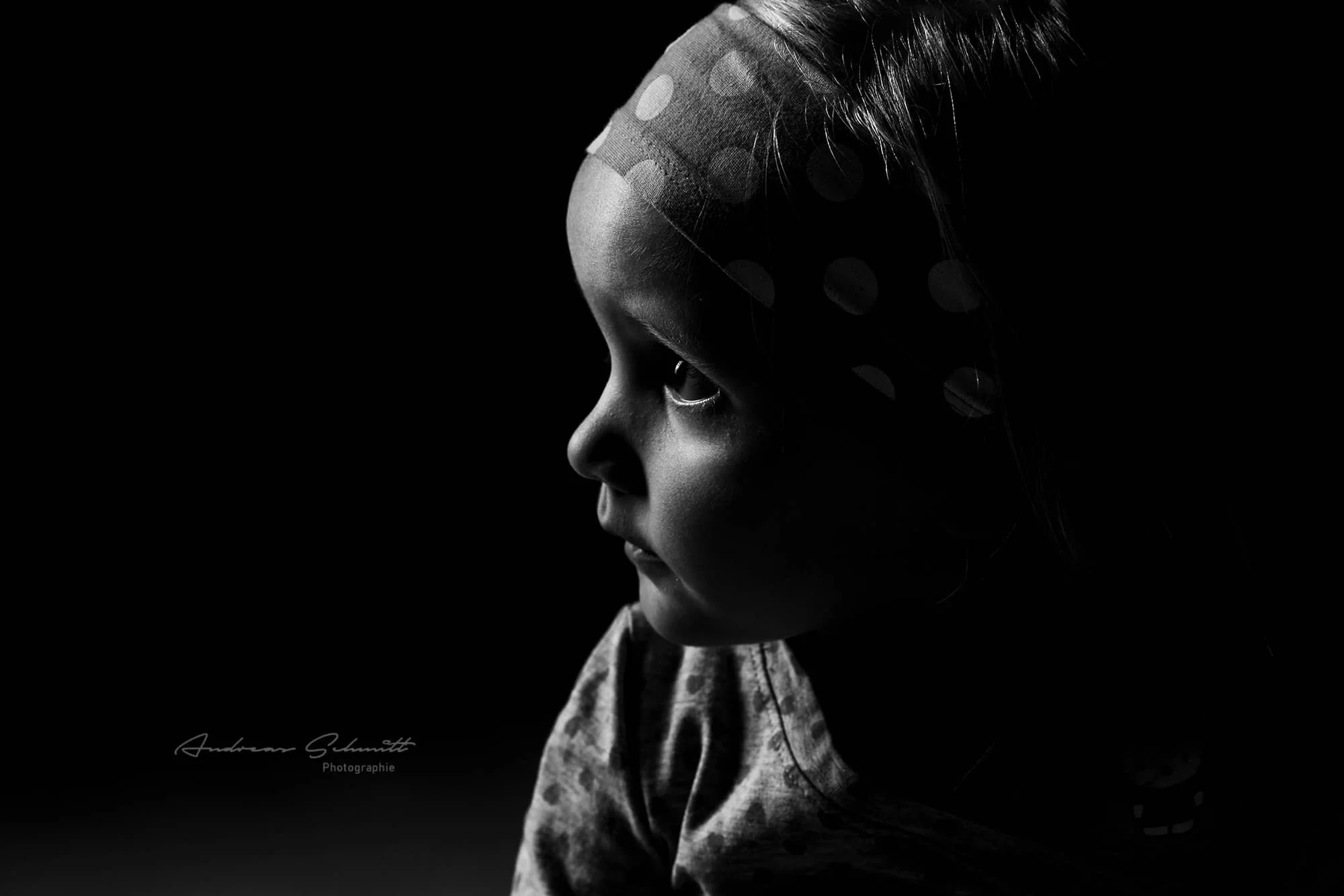 Schwarz-Weiß-Bild eines jungen Mädchens in Seitenprofil, das nach links schaut, mit Polkadot-Headband und casual Kleidung, dunkler Hintergrund.