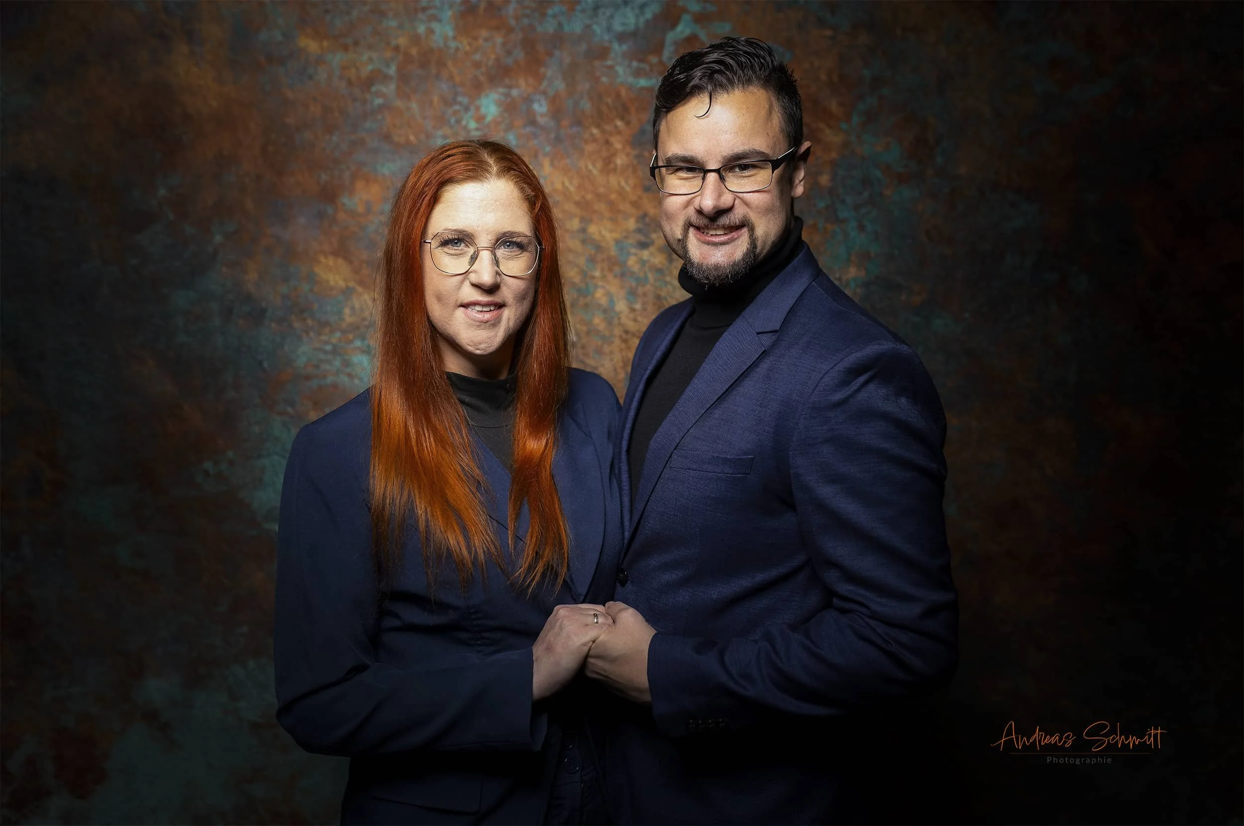 Ein Mann und eine Frau stehen sich nahe, halten die Hände und schauen in die Kamera, vor einem dunklen, kunstvoll gestalteten Hintergrund, beide tragen formelle dunkle Kleidung.