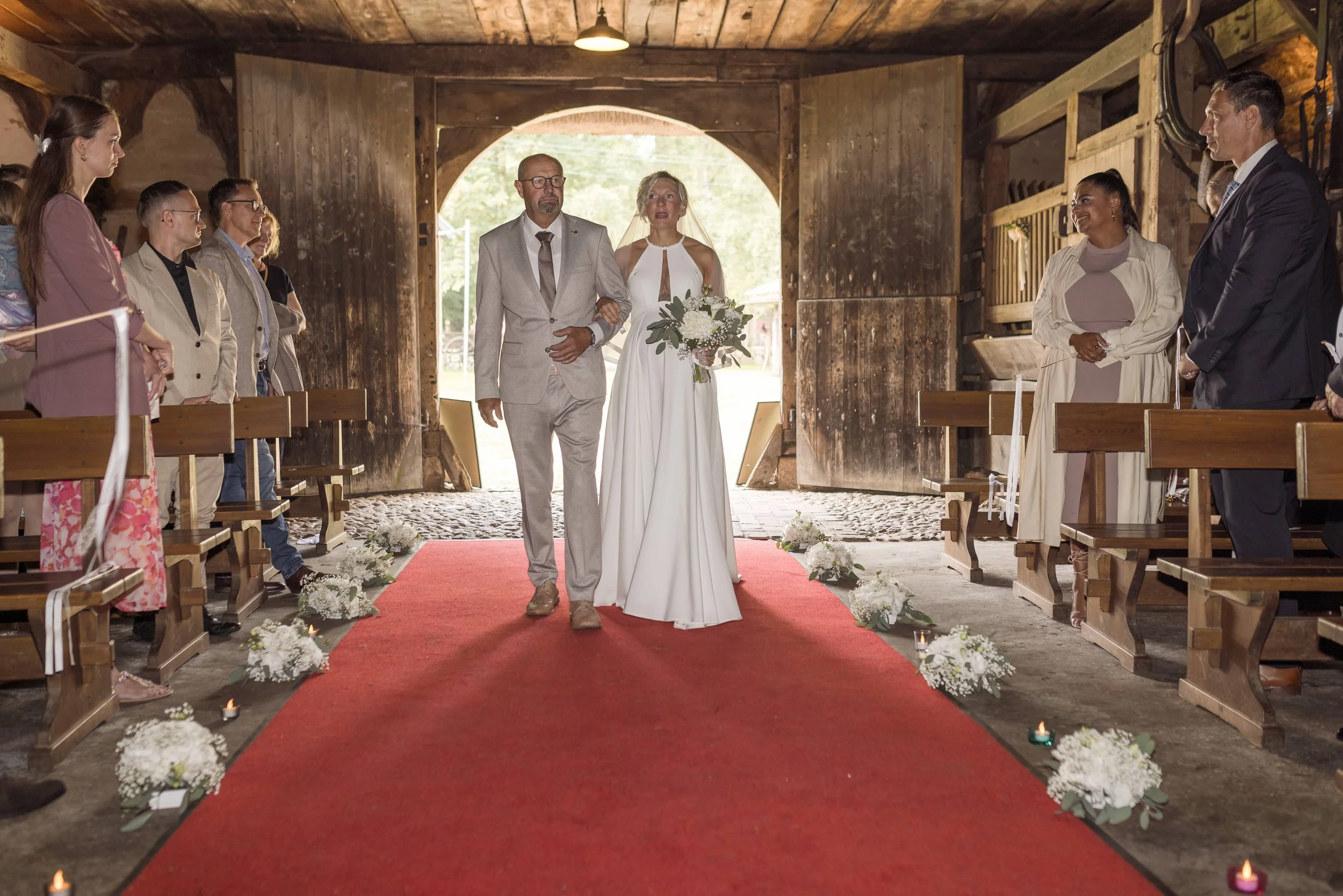 Braut wird vom Bräutigam in einer Scheune bei der Hochzeit begleitet, umgeben von Gästen, mit Blumen und Kerzen auf dem roten Teppich.