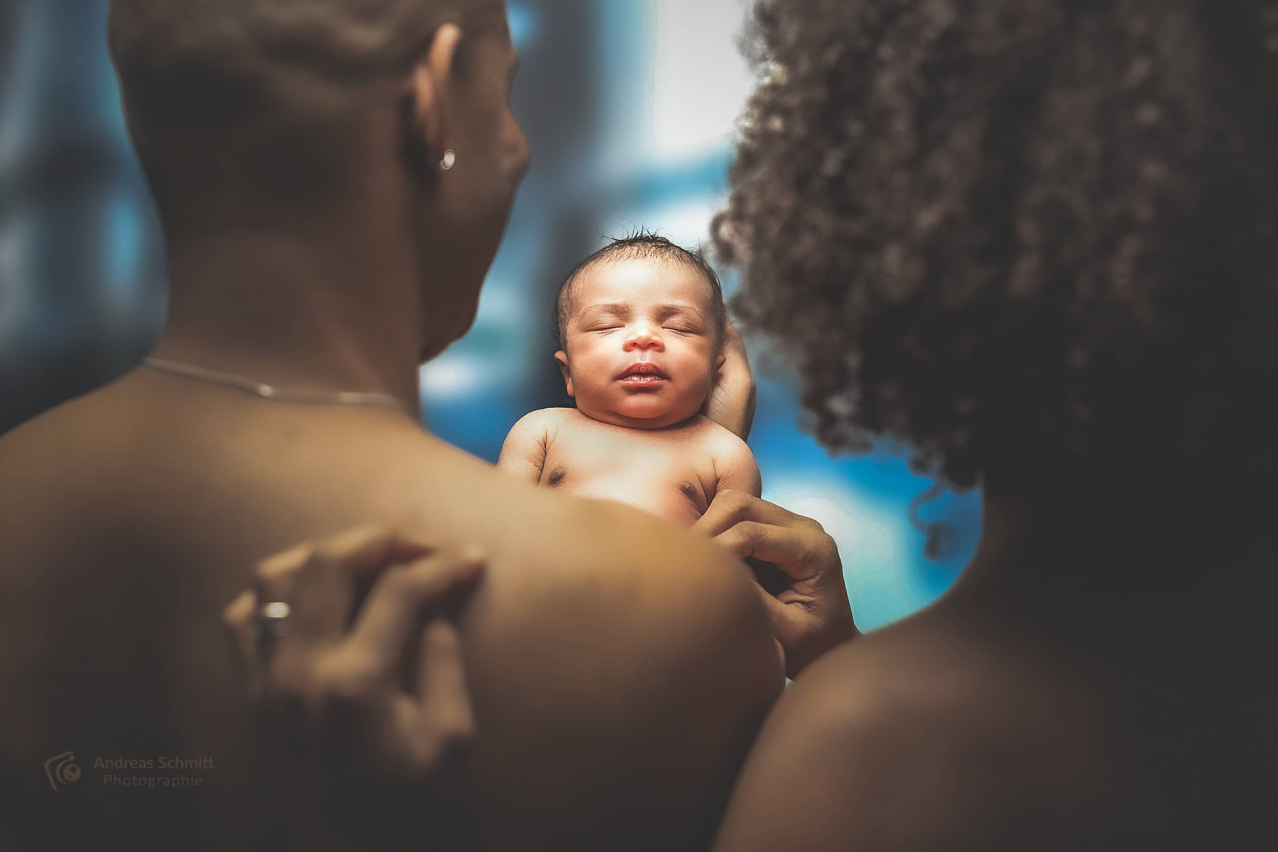 Eine schwangere Frau hält ein schlafendes Neugeborenes in den Armen, umgeben von einer anderen Person, die auf das Baby schaut, bei natürlichem Licht im Hintergrund.