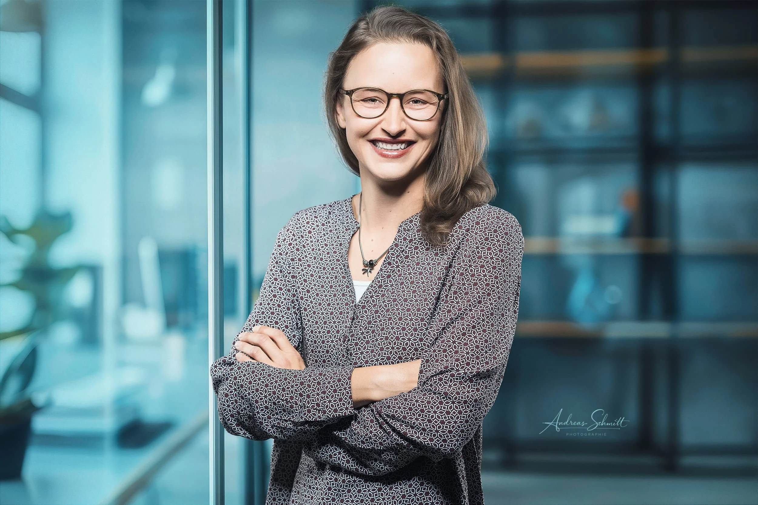 Lächelnde junge Frau mit Brille, Arme verschränkt, vor einer blauen modernen Büro-Umgebung.