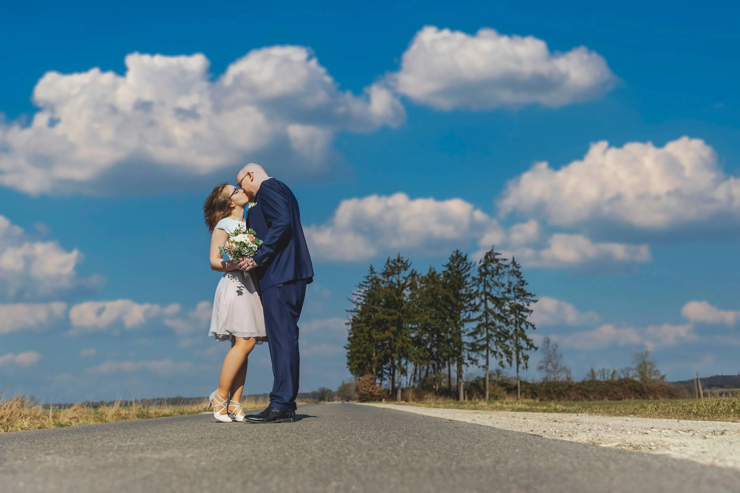 Ein Brautpaar auf einer Landstraße unter einem blauen Himmel mit Wolken, das sich küsst. Der Mann trägt einen blauen Anzug, die Frau ein weißes Kleid und hält einen Blumenstrauß.