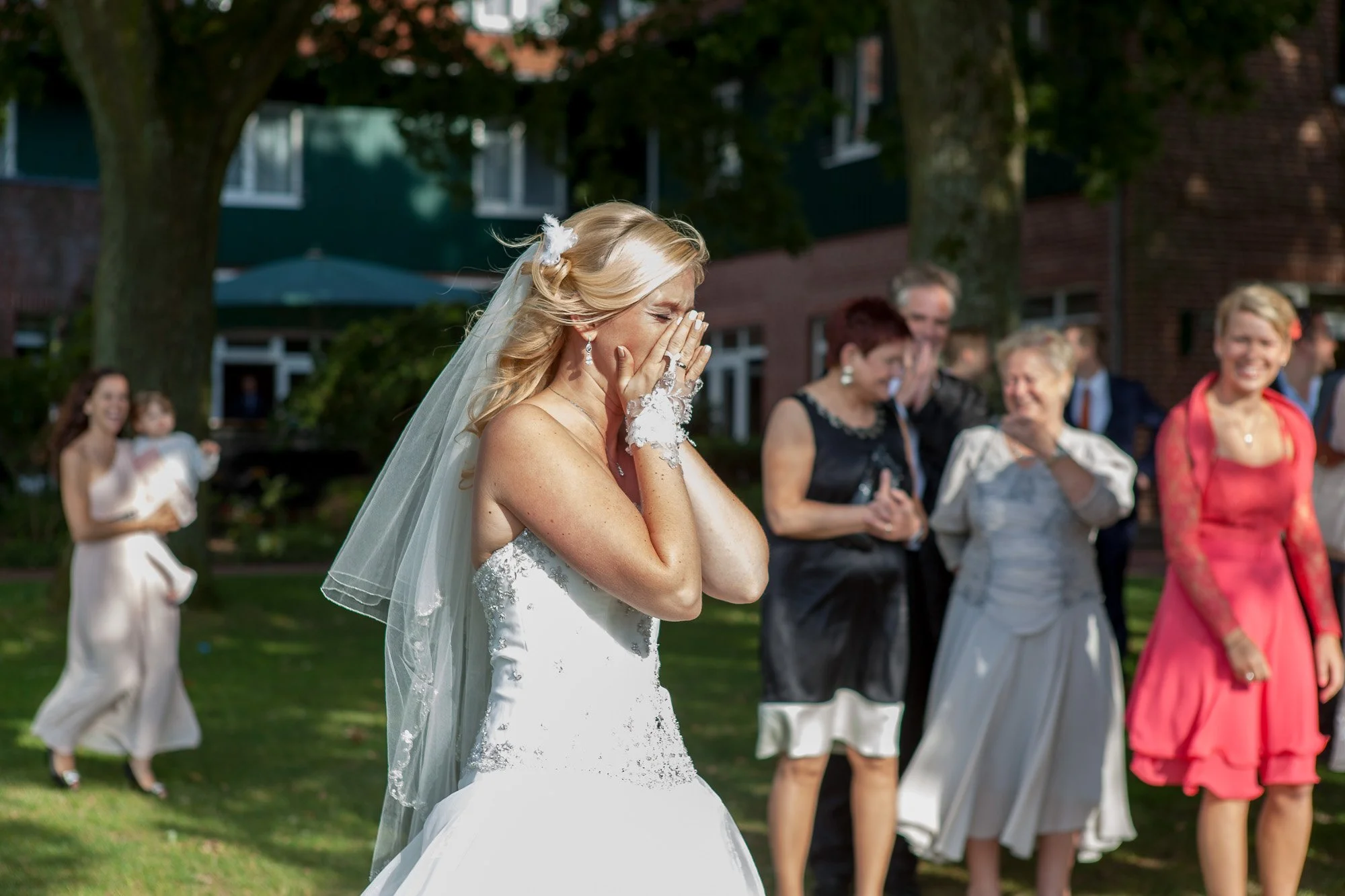 Eine emotionale Szene bei einer Hochzeit, eine Braut in einem weißen Kleid weint und bedeckt ihr Gesicht, während ihre Gäste im Hintergrund lachen und sich freuen.