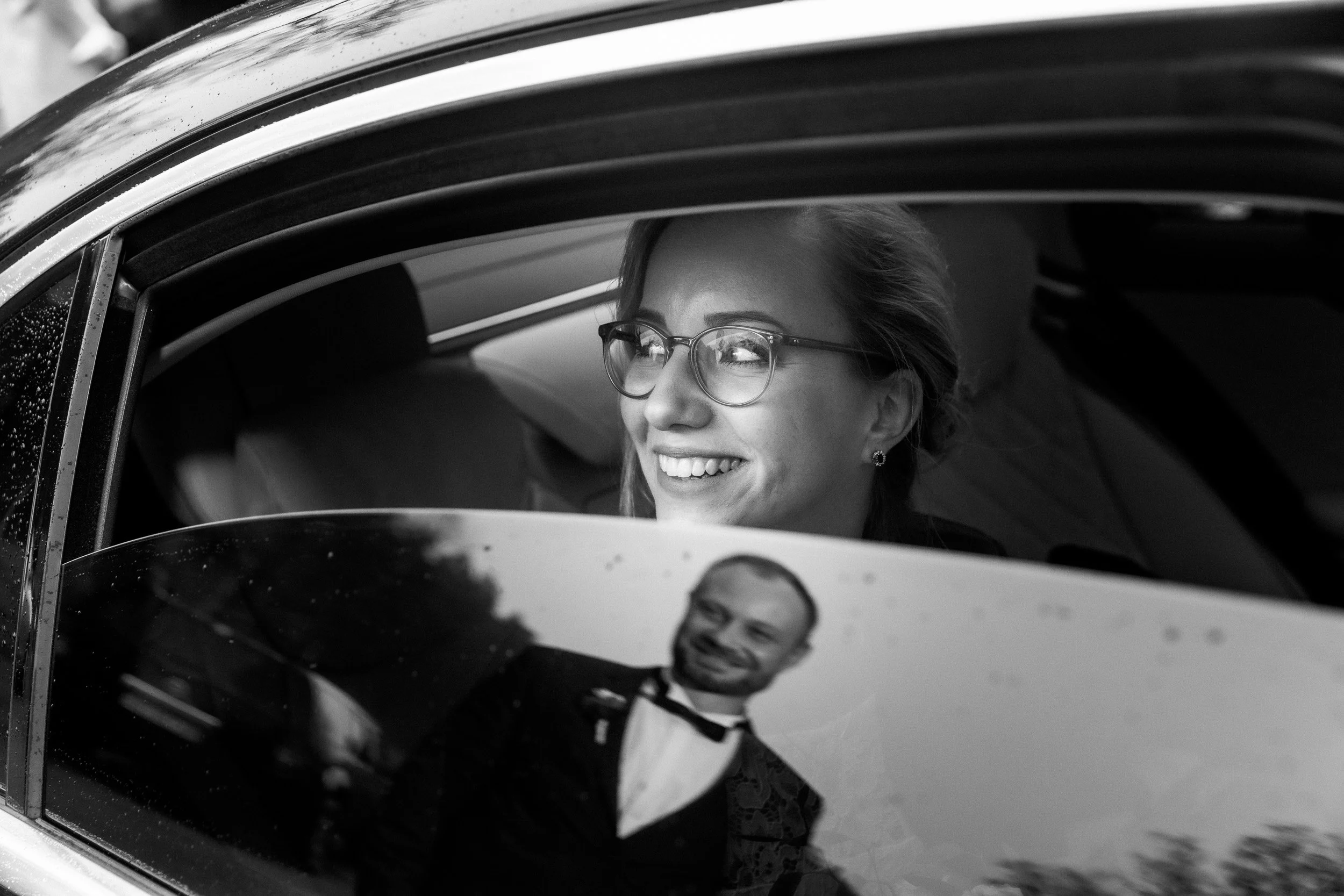 Ein lachendes Paar, vermutlich bei einer Hochzeit, sitzt in einem Auto. Die Frau trägt Brille und ist im Mittelpunkt, während der Mann in formeller Kleidung im Autofach zu sehen ist.
