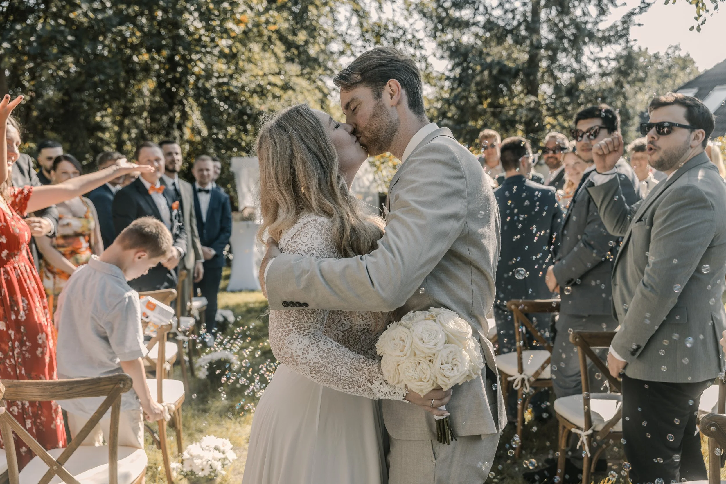 Ein Brautpaar küsst sich bei einer Außenehe im Sonnenlicht, umgeben von Gästen, die sich freuen und Bubbles blasen.