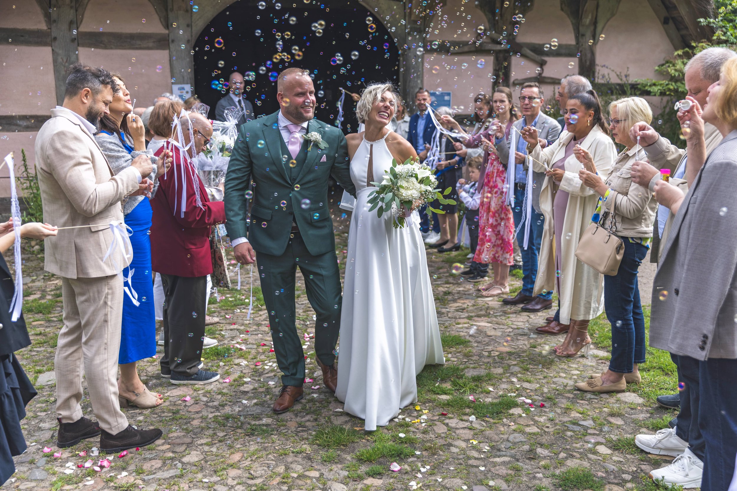 Ein glückliches Brautpaar wird bei einer Hochzeitsfeier von Gästen mit Seifenblasen umgeben, im Freien auf einer gepflasterten Fläche, mit einem historischen Gebäude im Hintergrund.