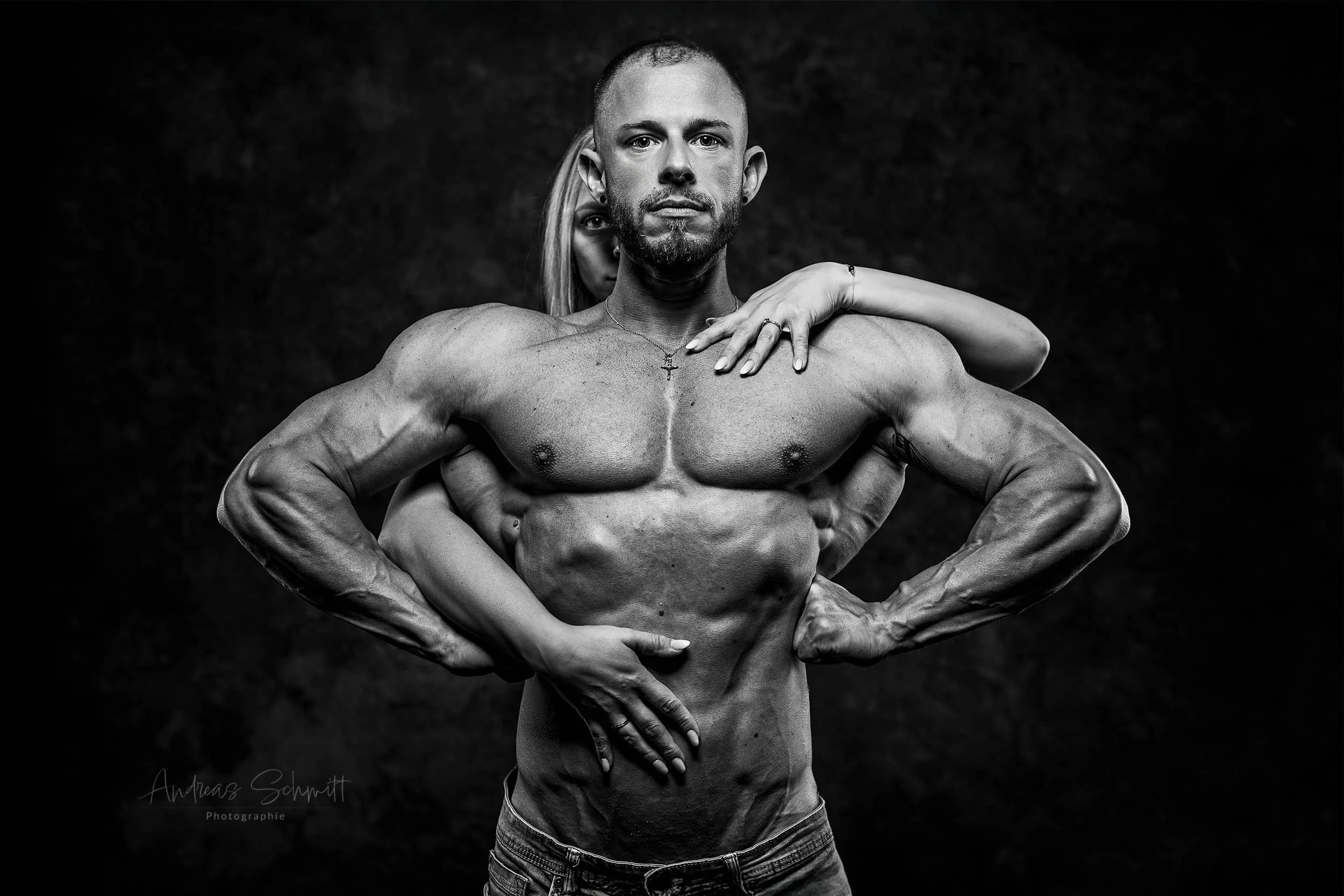 Schwarze und weiße Fotografie eines muskulösen Mannes mit kurzen Haaren und einem kleinen Kreuz-Halskette, mit einer Person hinter ihm, deren Arme ihn umarmen, alle vor dunklem Hintergrund.