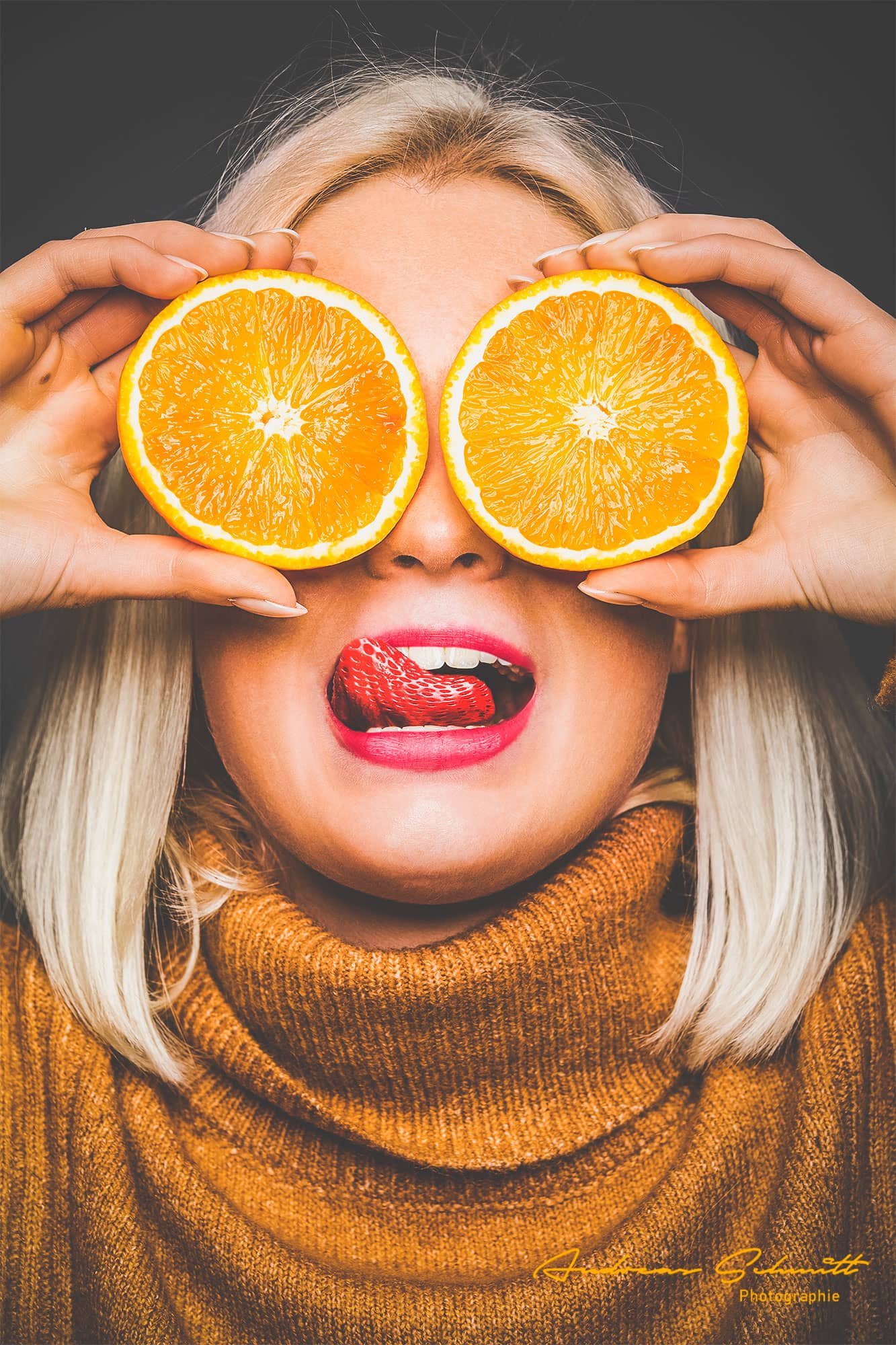 Frau mit blonden Haaren hält zwei Orangenscheiben vor die Augen, im Mund eine Erdbeere, trägt roten Lippenstift und einen braunen Pullover.