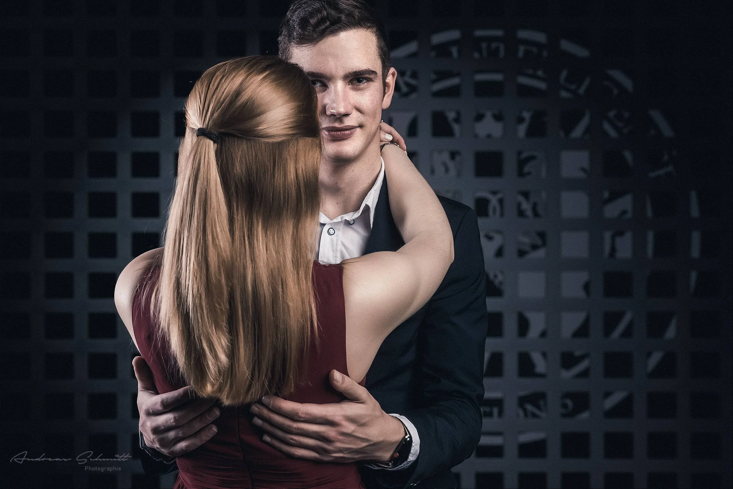 Ein Mann trägt einen schwarzen Anzug und eine weiße Hemd, während eine Frau mit langen roten Haaren sie umarmt und ihr Gesicht an seiner Wange versteckt, vor einem dunklen Hintergrund.