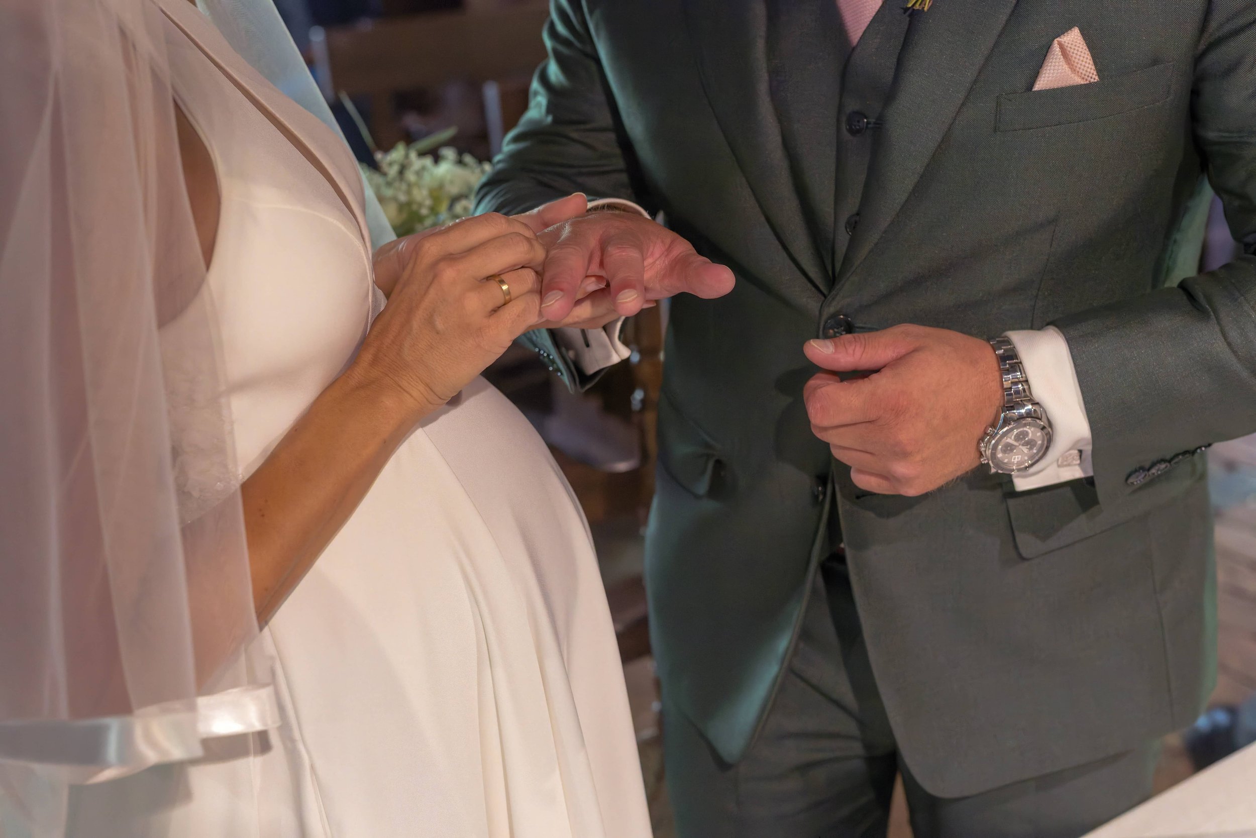Hände eines Paares beim Heiratsritual, ein Mann legt einen Ring an die Hand einer Frau, im Hintergrund ist eine Hochzeitsszene sichtbar.