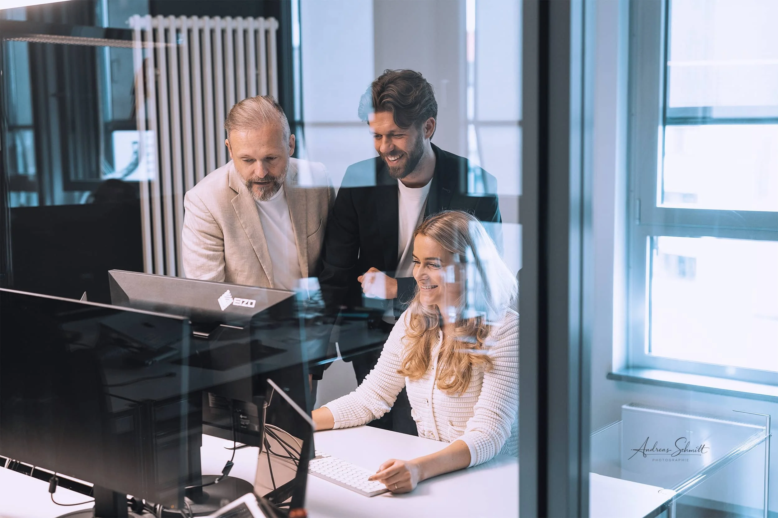 Zwei Männer und eine junge Frau schauen auf einen Computerbildschirm in einem modernen Büro, während sie zusammenarbeiten und lachen.