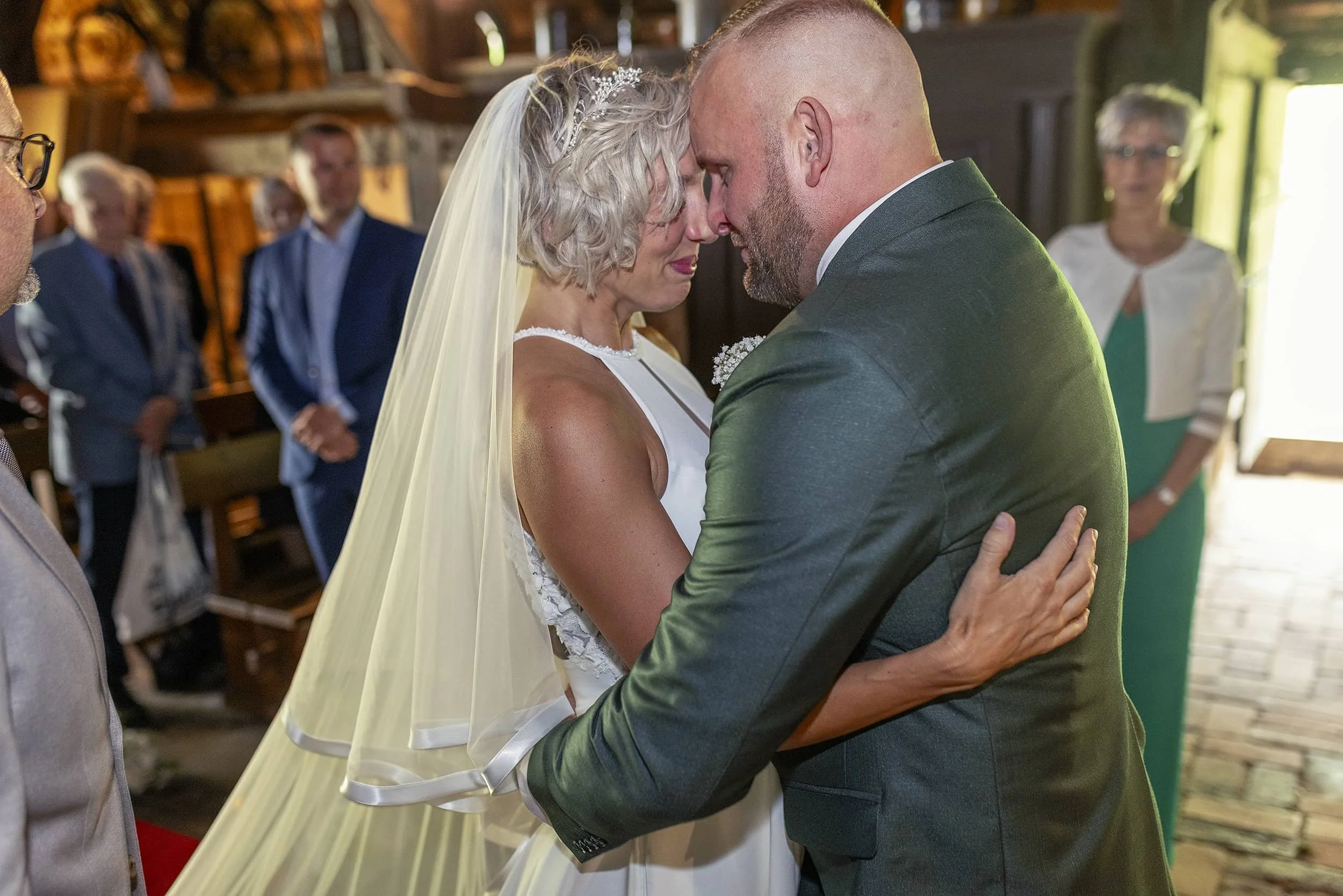 Ein Brautpaar beim Heiratsgelübde, die Braut trägt ein weißes Hochzeitskleid mit Schleier und der Bräutigam einen dunklen Anzug, in einer Kirche.
