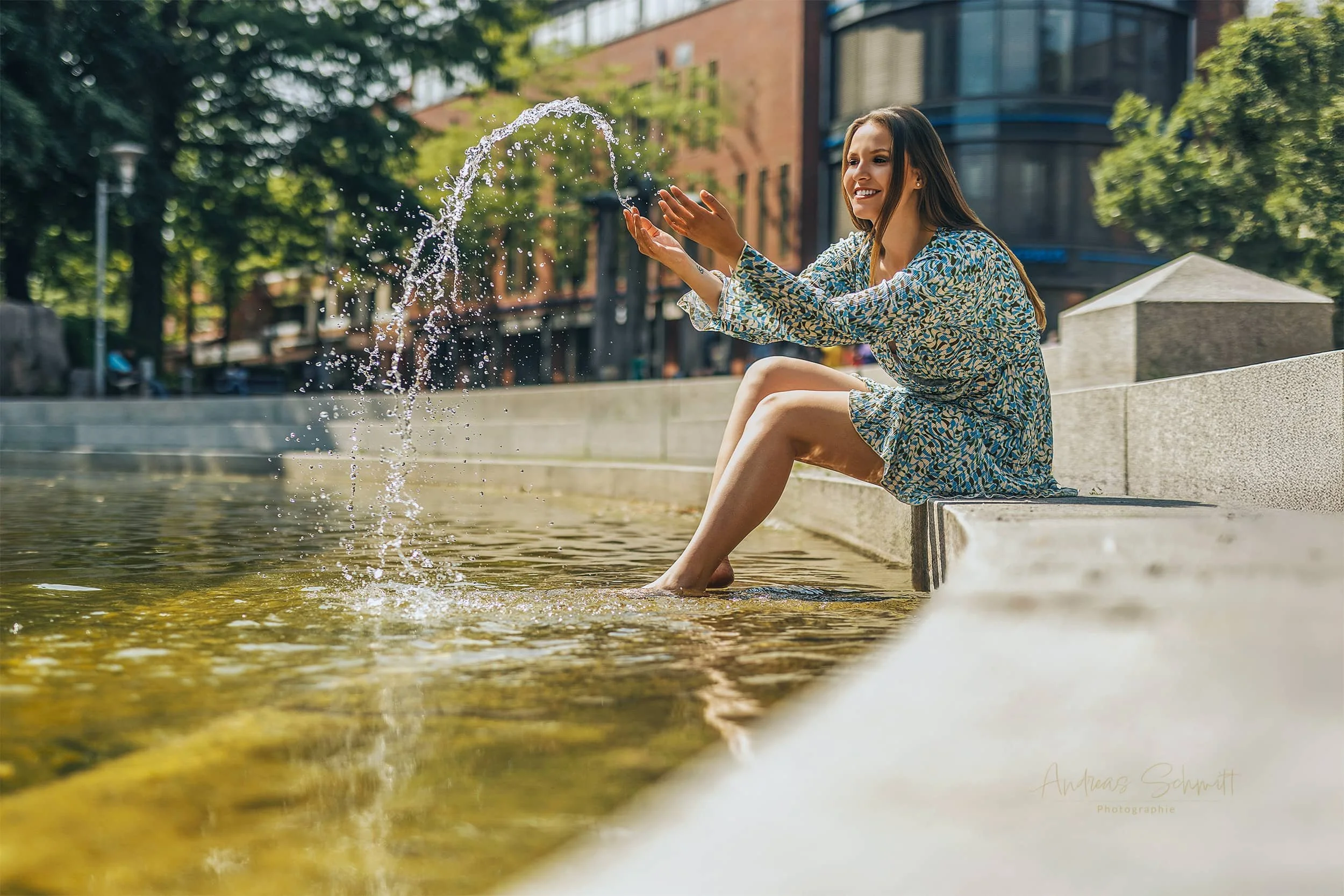 eine Frau sitzt im Sommer in einem Sommerkleid barfuß an einem Wasserbecken und spritzt fröhlich mit Wasser