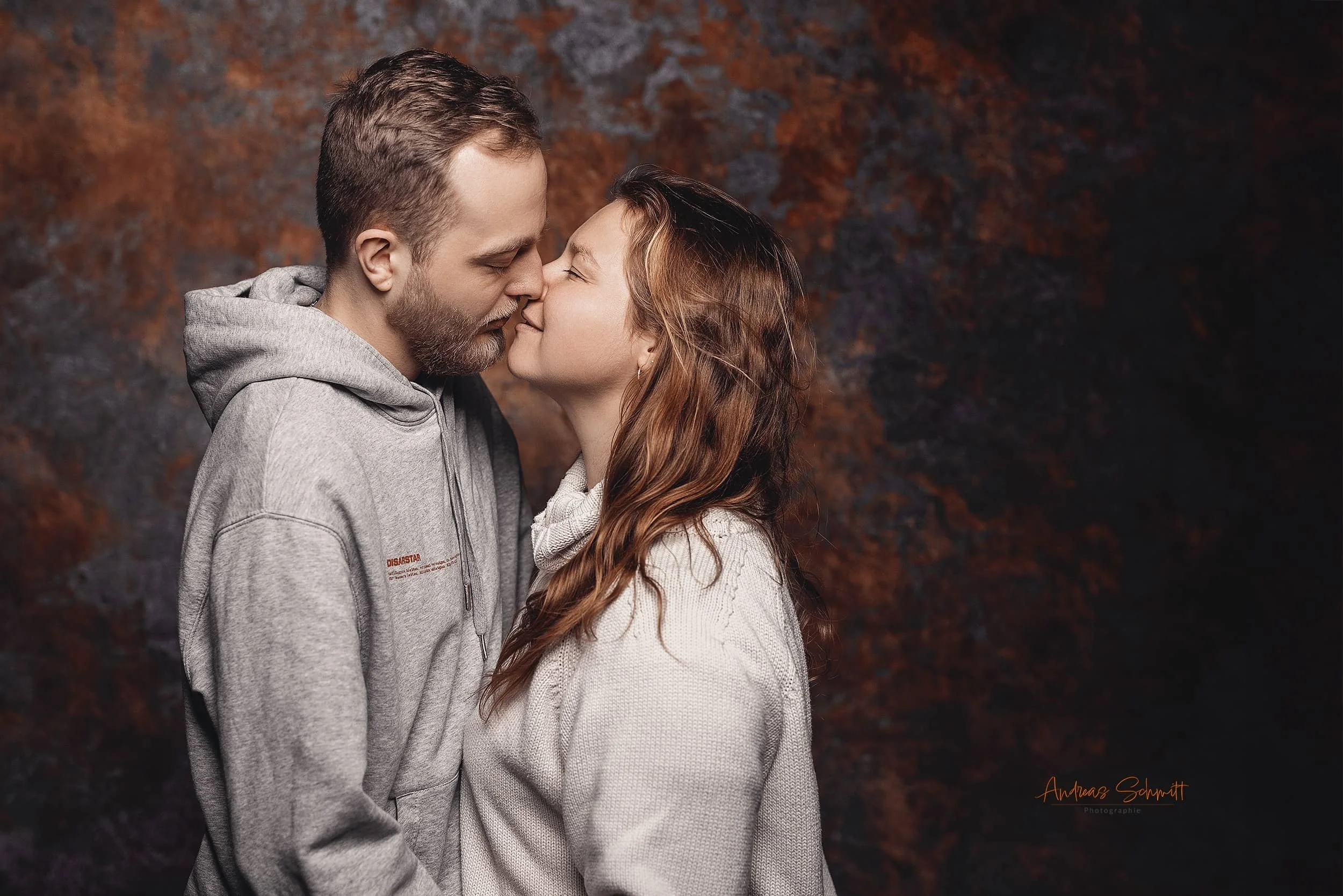 Ein Paar steht sich nahe gegenüber, mit abgesenkten Köpfen und den Nasen sich berührend, vor einem dunklen, rötlichen Hintergrund.