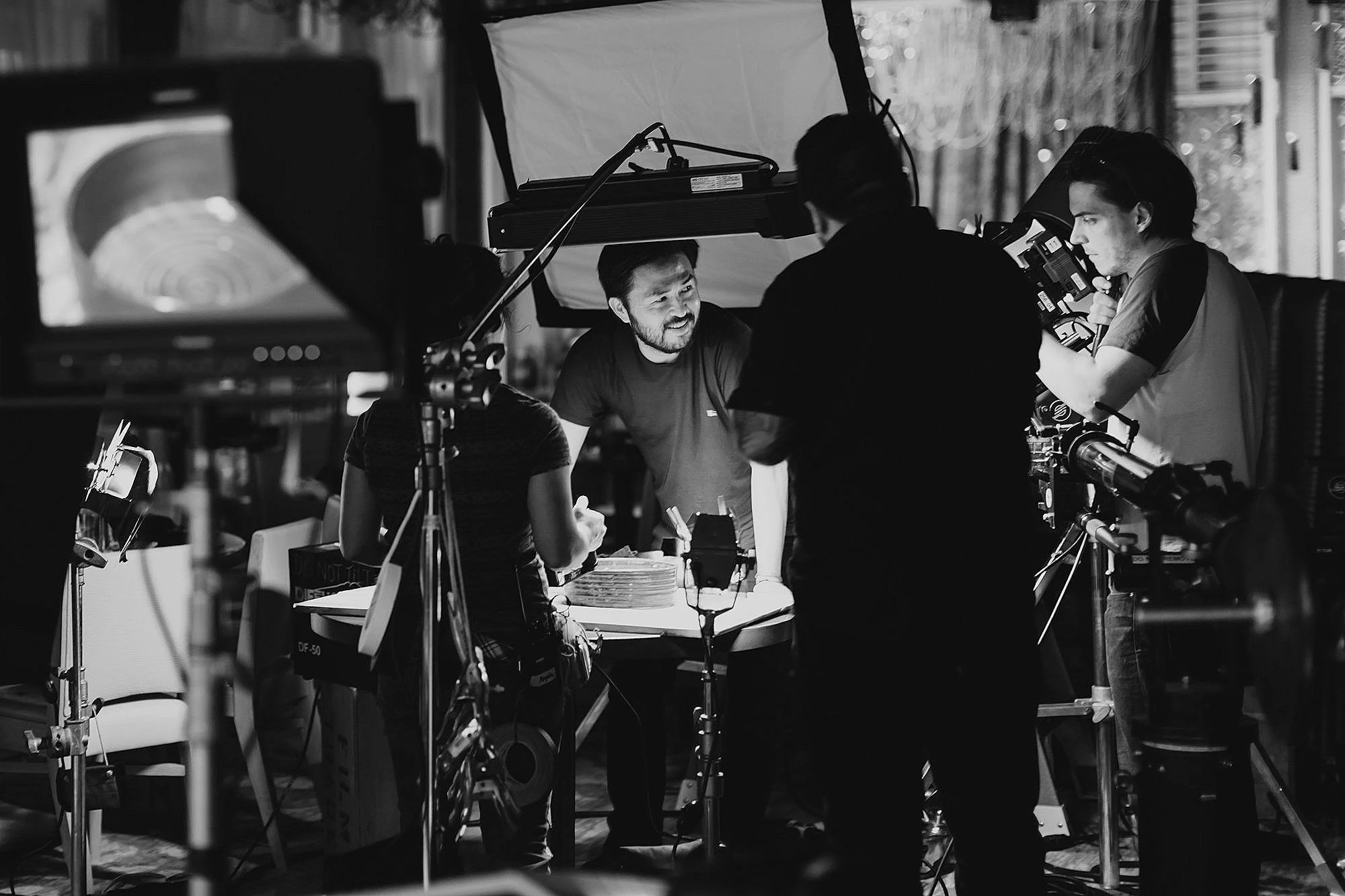 Filming crew working on a video shoot, with a man smiling at the center, surrounded by equipment and camera operators in an indoor setting.