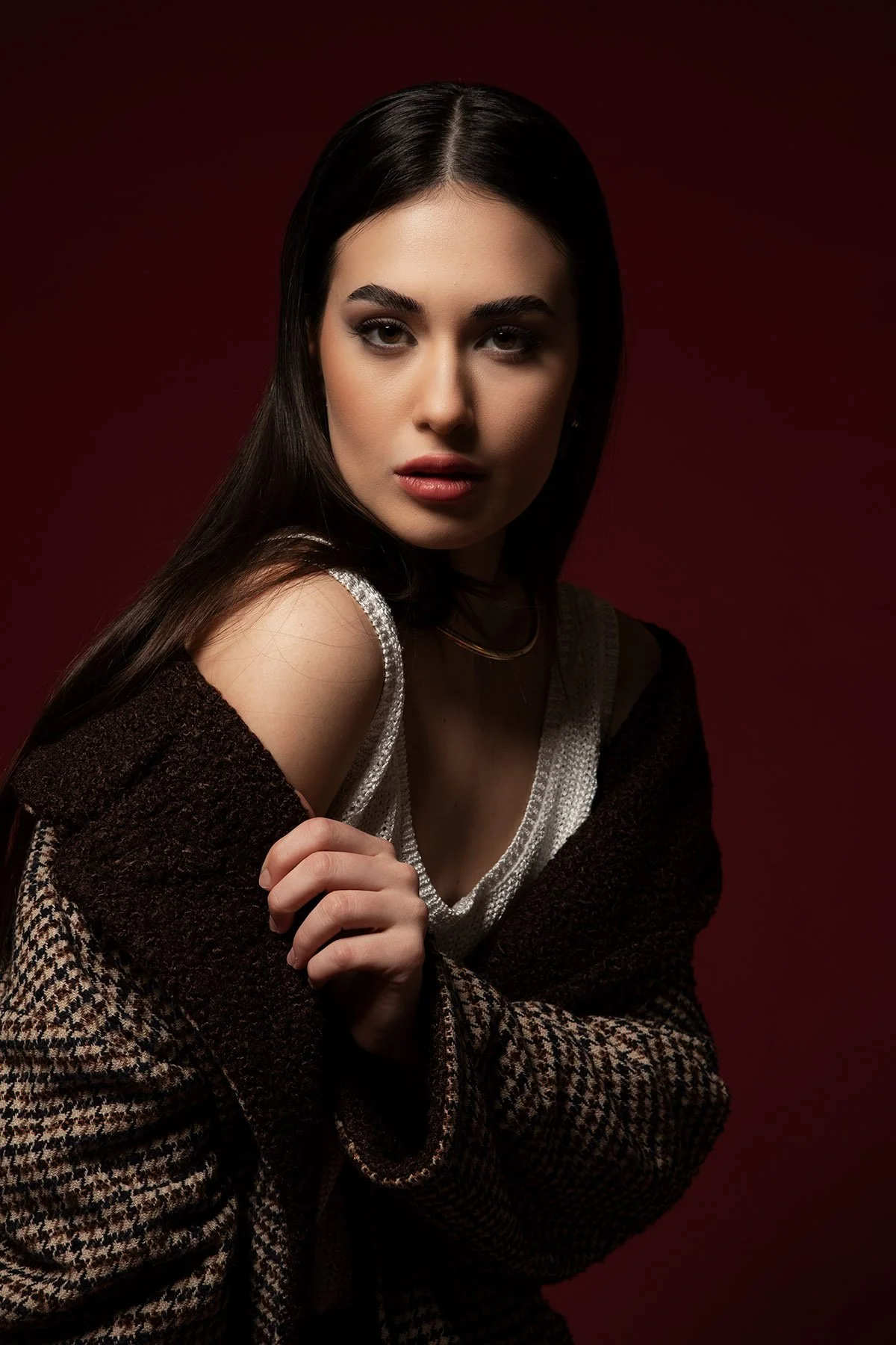 Ragazza con capelli scuri e trucco elegante, indossa un abito bianco e una giacca scozzese, sfondo rosso scuro.