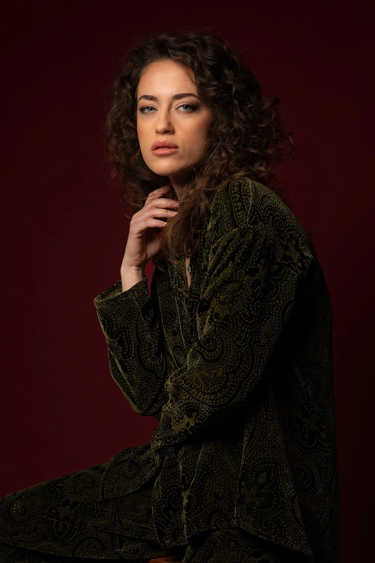 Ritratto di una donna con capelli ricci e occhi azzurri, indossa un abito scuro con un ricamo complesso, sullo sfondo di colore rosso scuro.