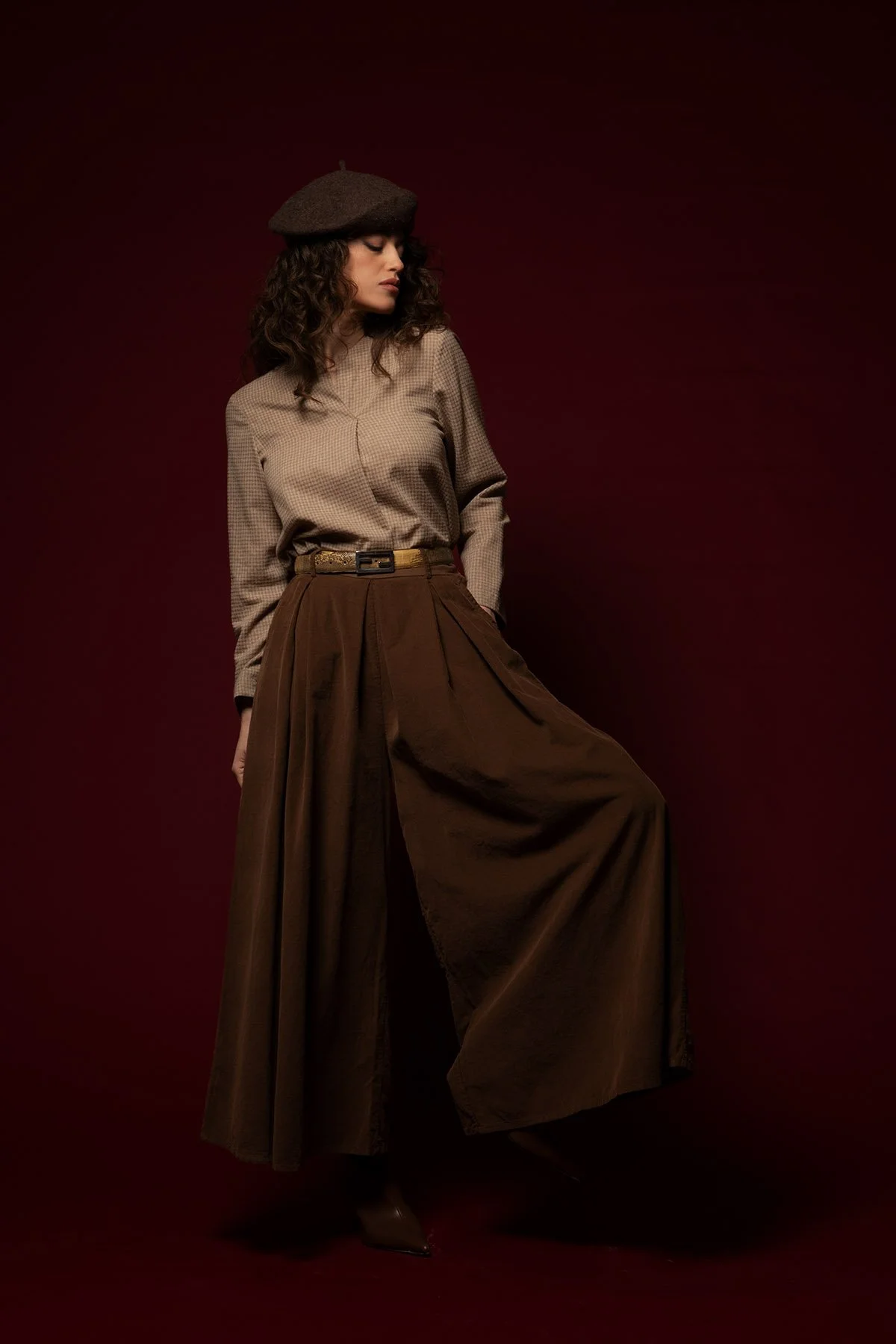 Donna con cappello brown, camicia a quadretti beige, e gonna a campana marrone scuro, posando contro sfondo rosso scuro.