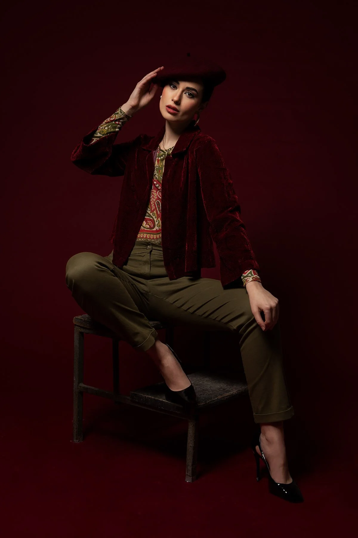 Donna seduta su una sedia, vestita con un cappotto di velluto rosso, una maglietta con motivo paisley e pantaloni kaki, con un cappello rosso. Sfondo rosso scuro.